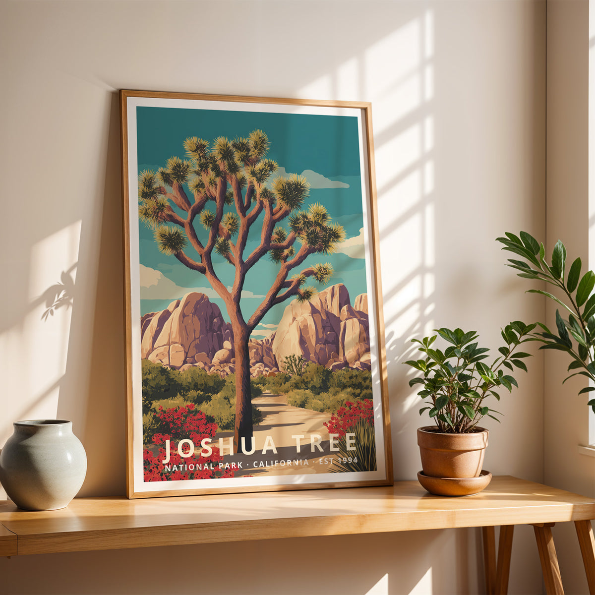 Joshua Tree National Park travel poster in a light oak frame on a wooden console table with potted plants in sunlight