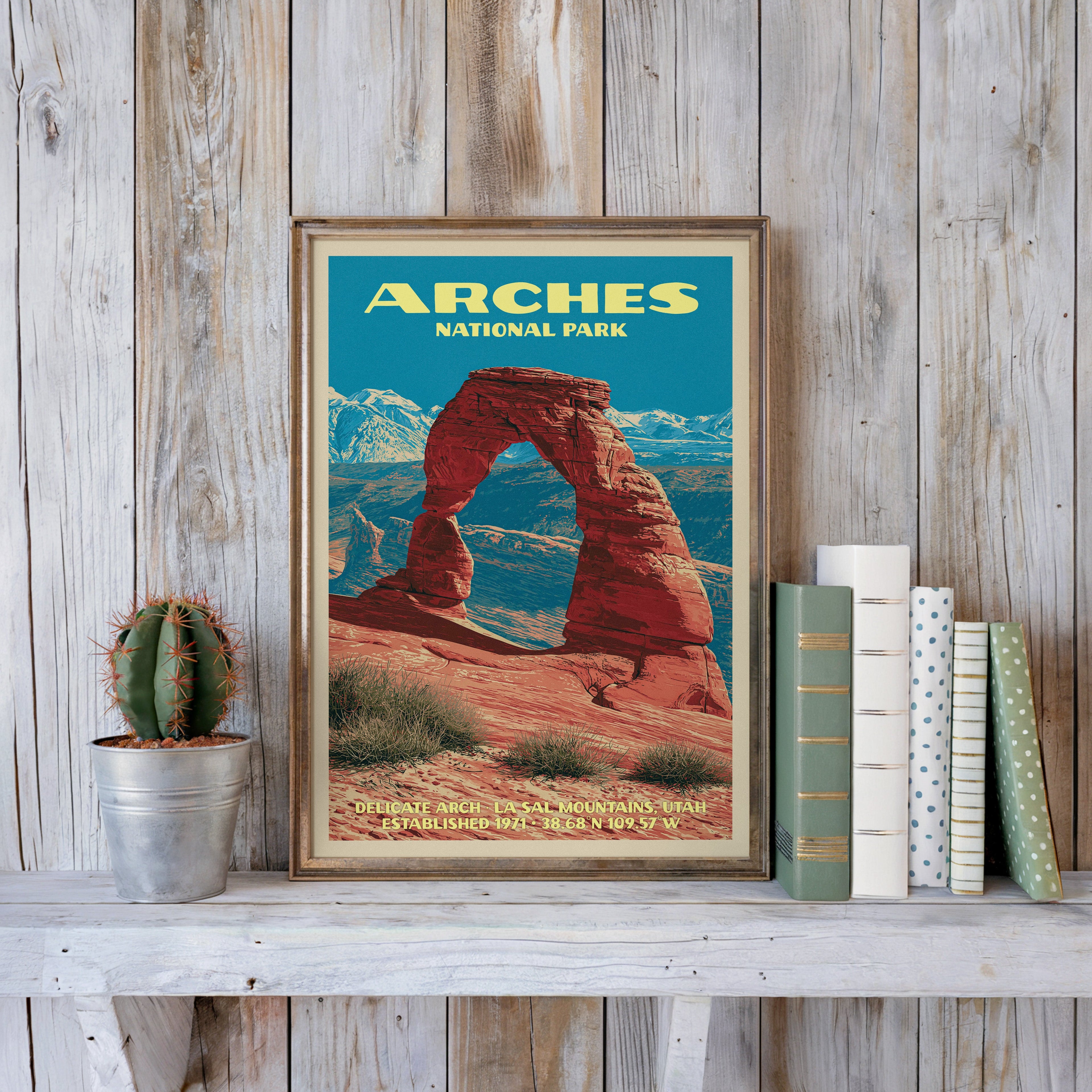 Arches vintage poster in a slim gold frame on a whitewashed plank wall beside a cactus and pastel books