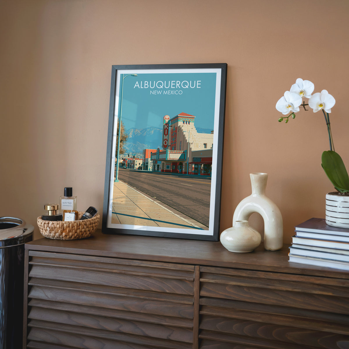 Central Avenue print, black frame on a walnut sideboard with terracotta wall, white orchid and cream ceramic vase.
