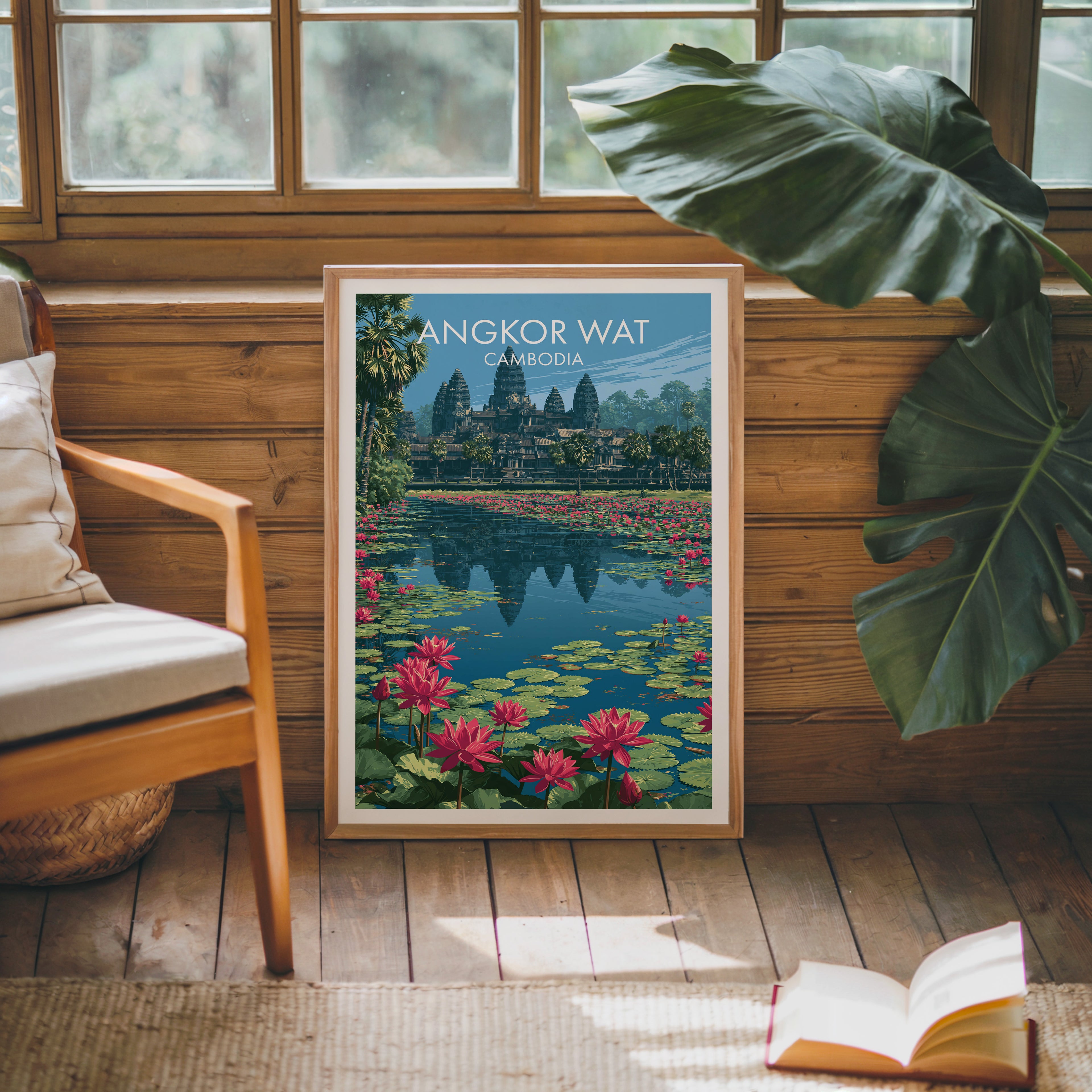 Framed Angkor Wat Cambodia poster leaning against a timber cabin wall with monstera leaves and an open book