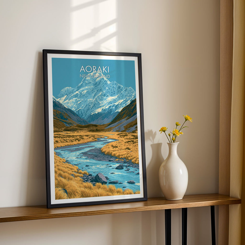 Snow-capped Aoraki artwork, black frame on a wood shelf with yellow daisies against a sunlit cream wall