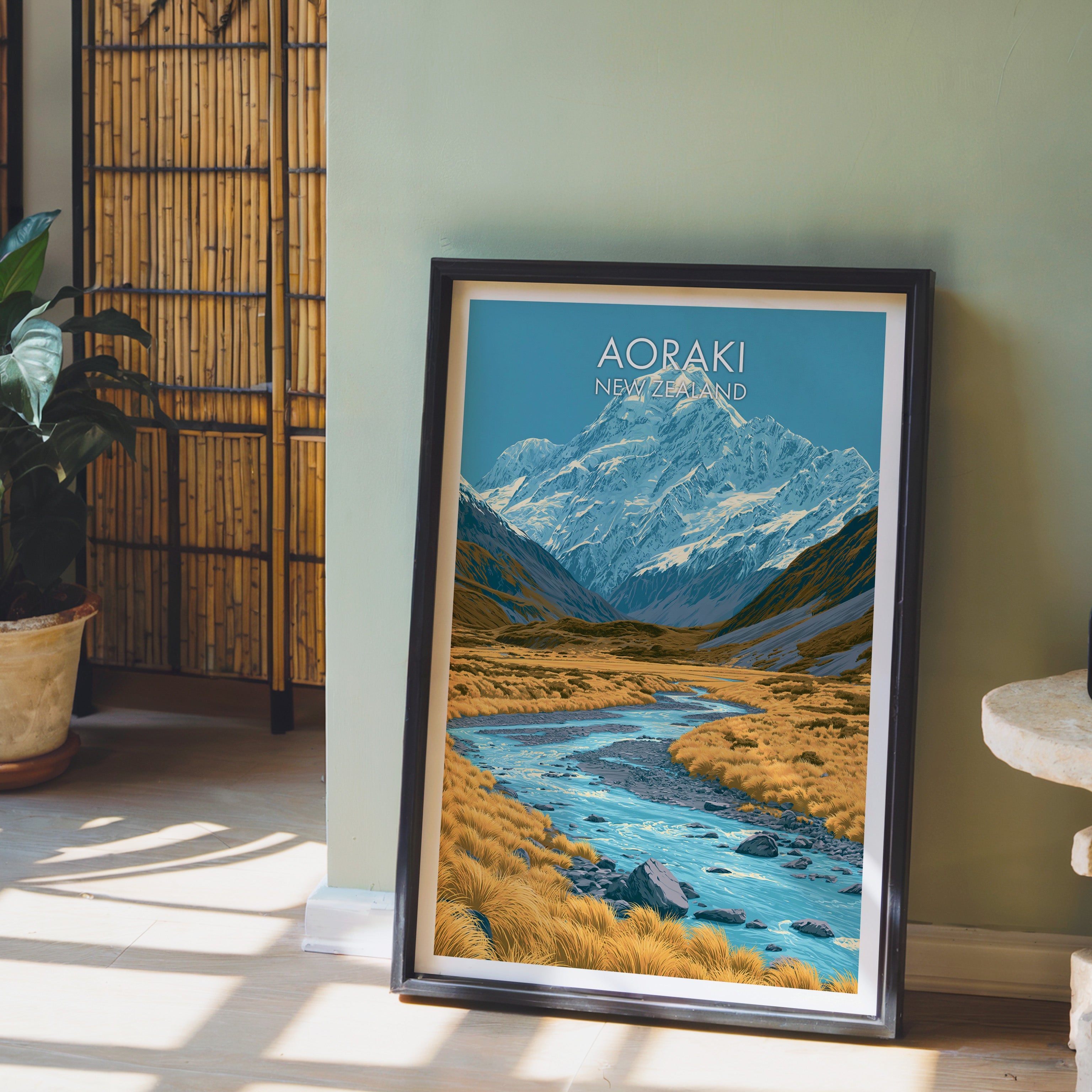 New Zealand mountain print in a black frame on a pale wood floor by a sage-green wall and bamboo screen
