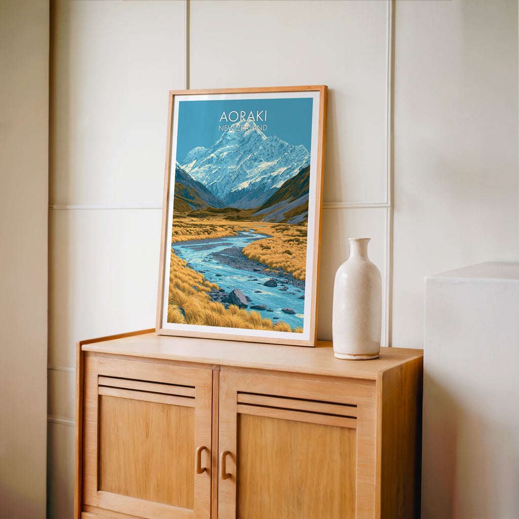 Southern Alps travel poster in an oak frame on a light oak cabinet beside a pale bottle vase