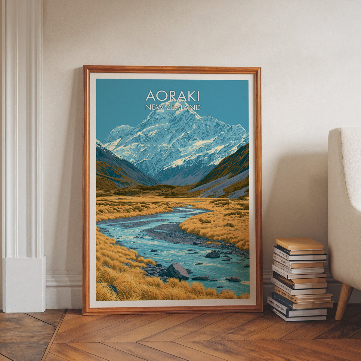 Aoraki artwork in a walnut frame on herringbone floor beside stacked books and a pale armchair