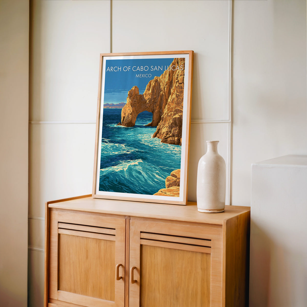 Coastal rock artwork in an oak frame on a light oak cabinet against a cream panelled wall, tall vase