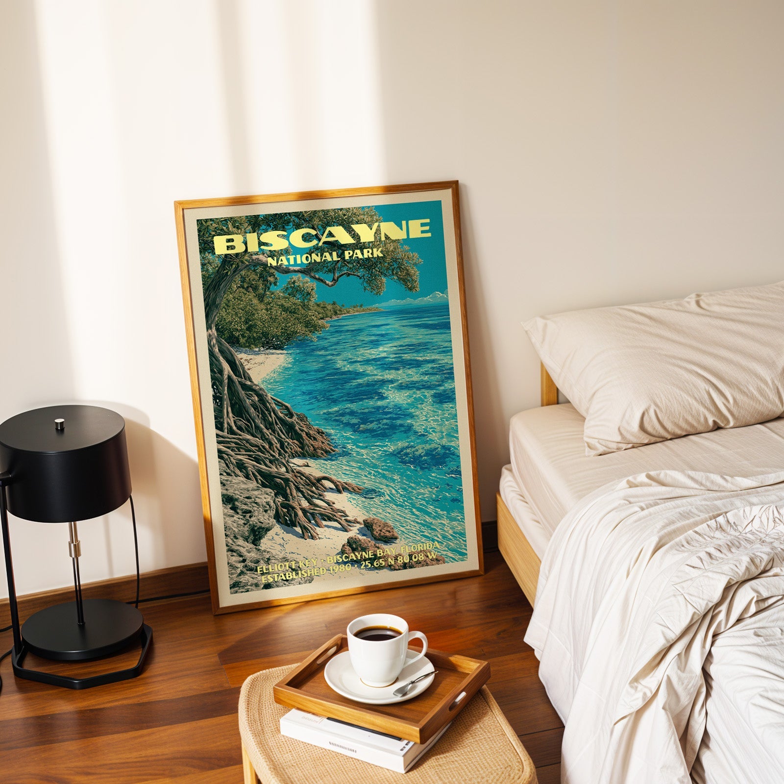 Elliott Key Biscayne poster in a gold frame on the floor beside a made bed and a black table lamp