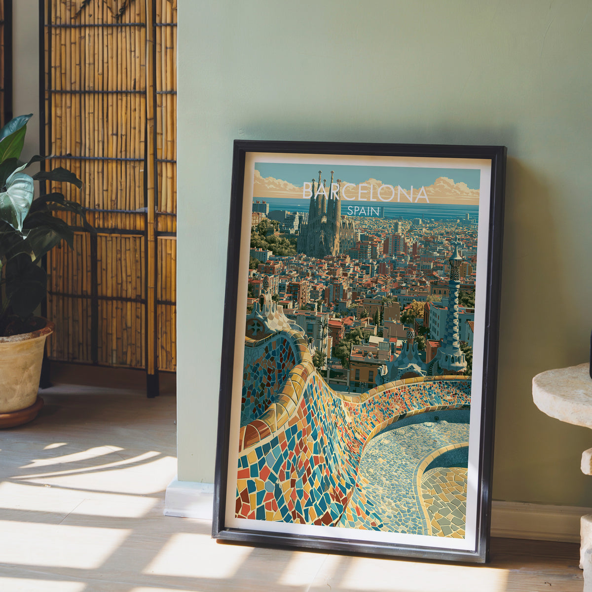 Barcelona Park Guell poster leaning on a sage green wall beside a bamboo screen and a potted plant in sunlight