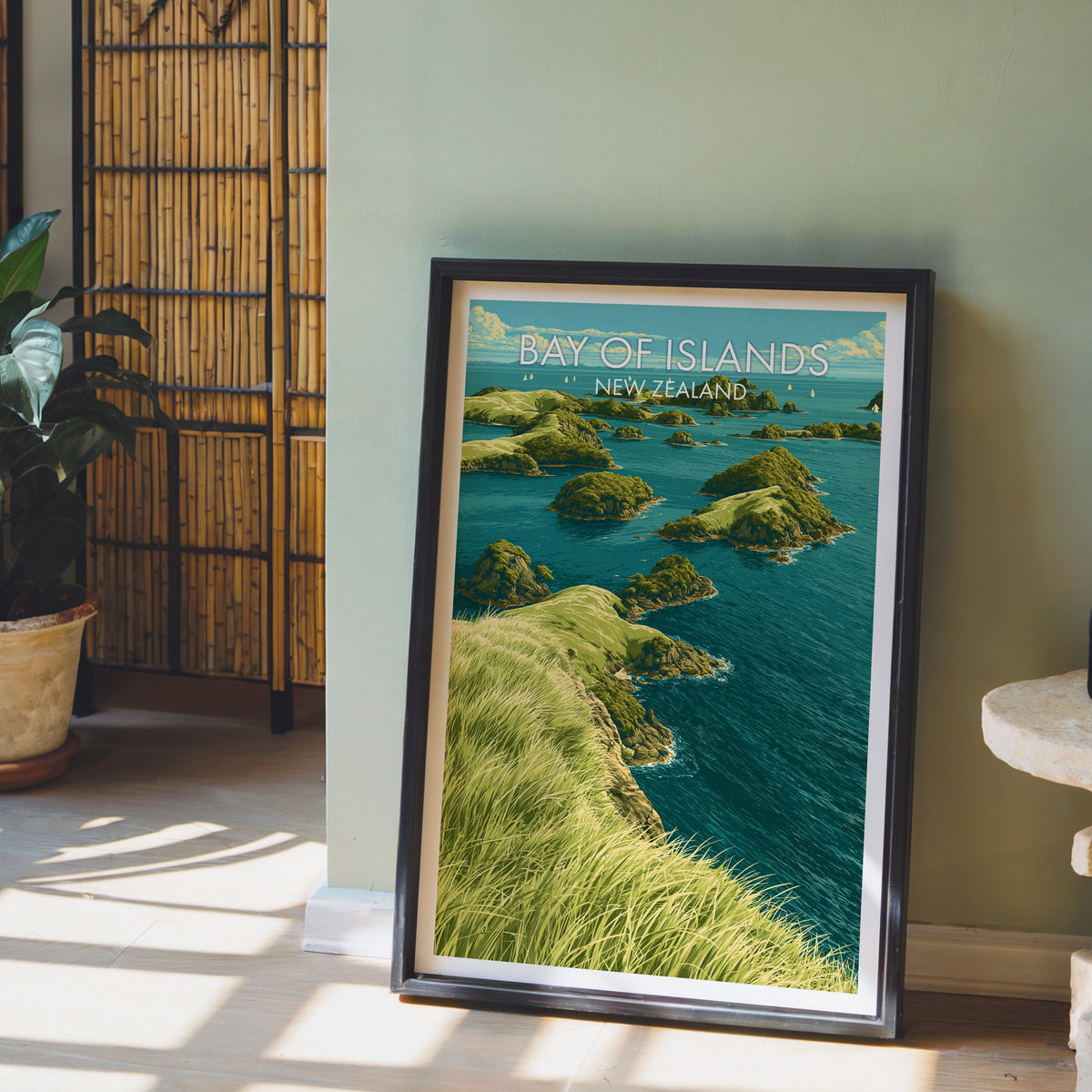 New Zealand island bay artwork in a black frame leaning on a pale wood floor by a sage-green wall and bamboo screen
