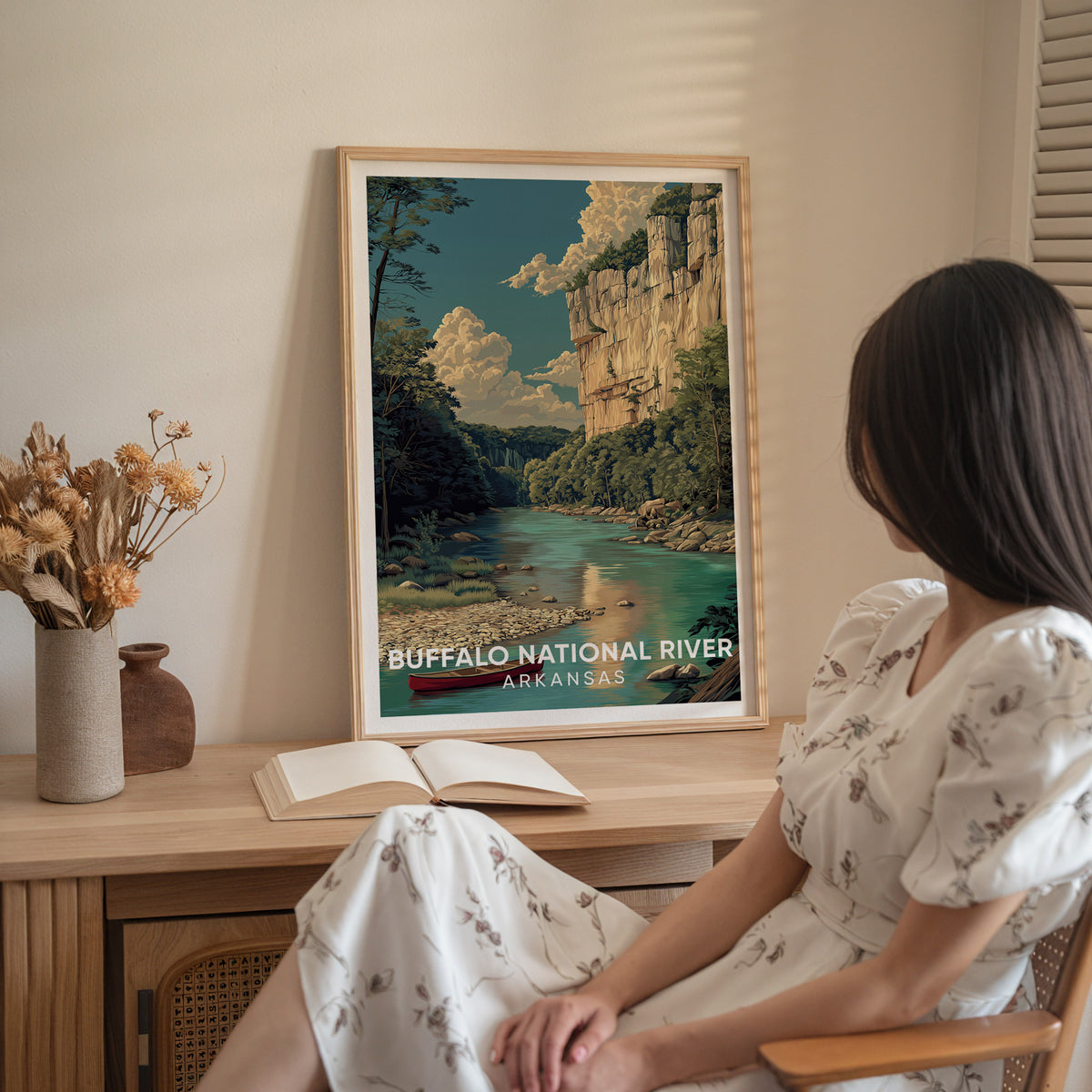 Woman in a white dress seated by an oak-framed Buffalo National River print and dried flowers