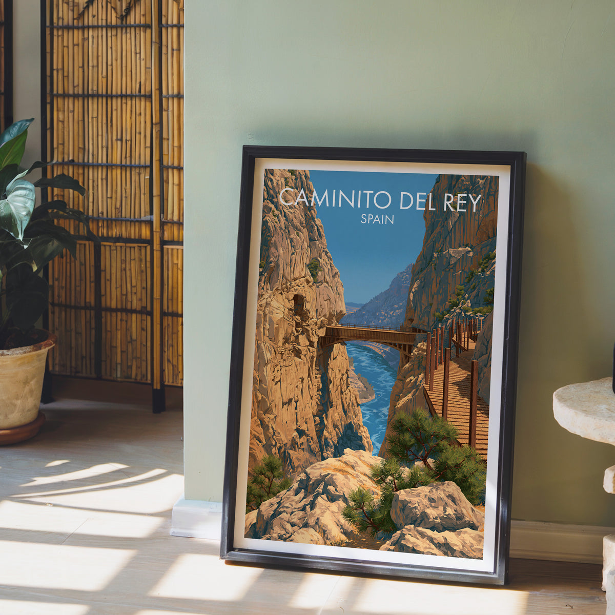 El Chorro gorge poster, dark frame leaning on a sage-green wall beside a bamboo screen and a peace lily