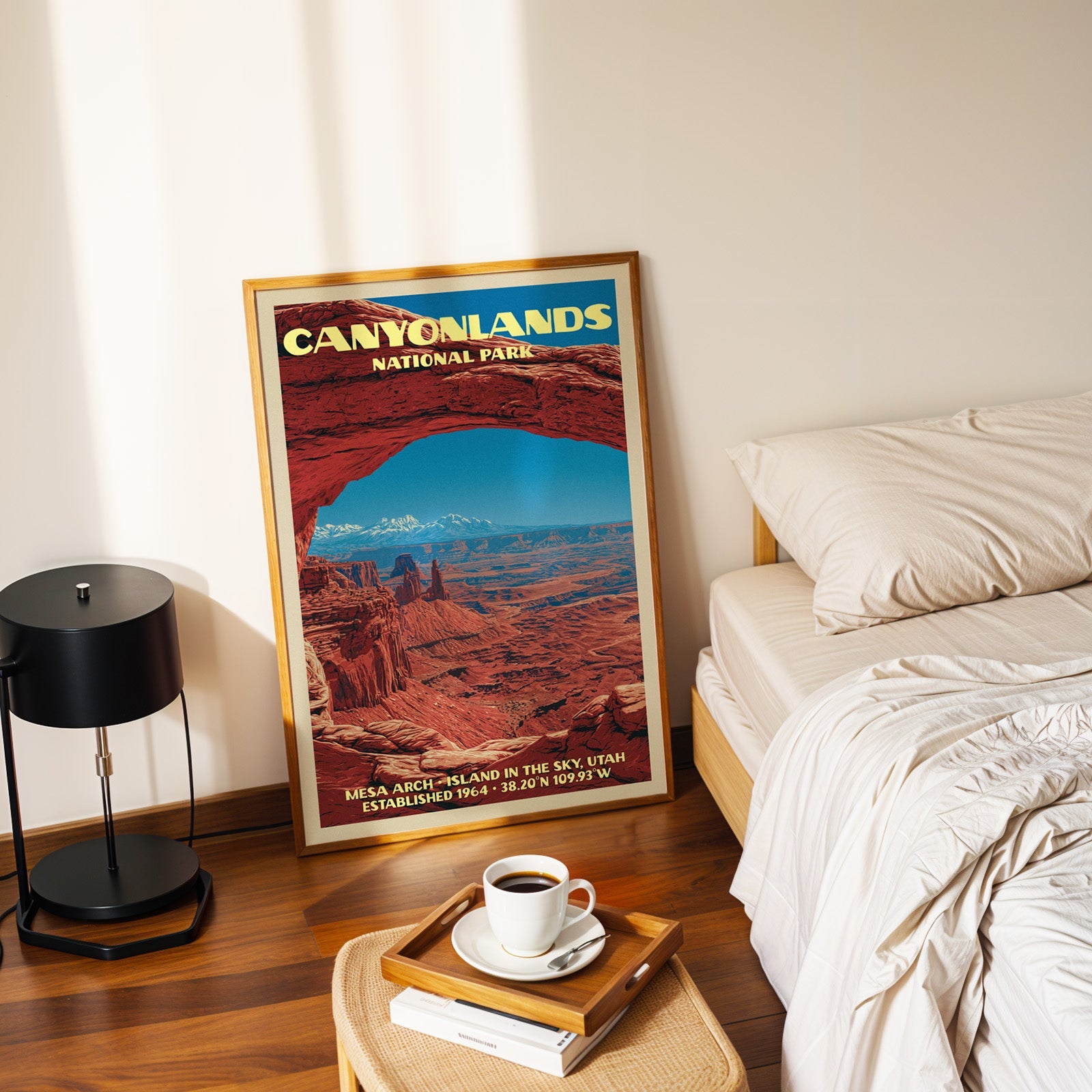 Mesa Arch Canyonlands poster in a gold frame on the floor beside a made bed and a black table lamp
