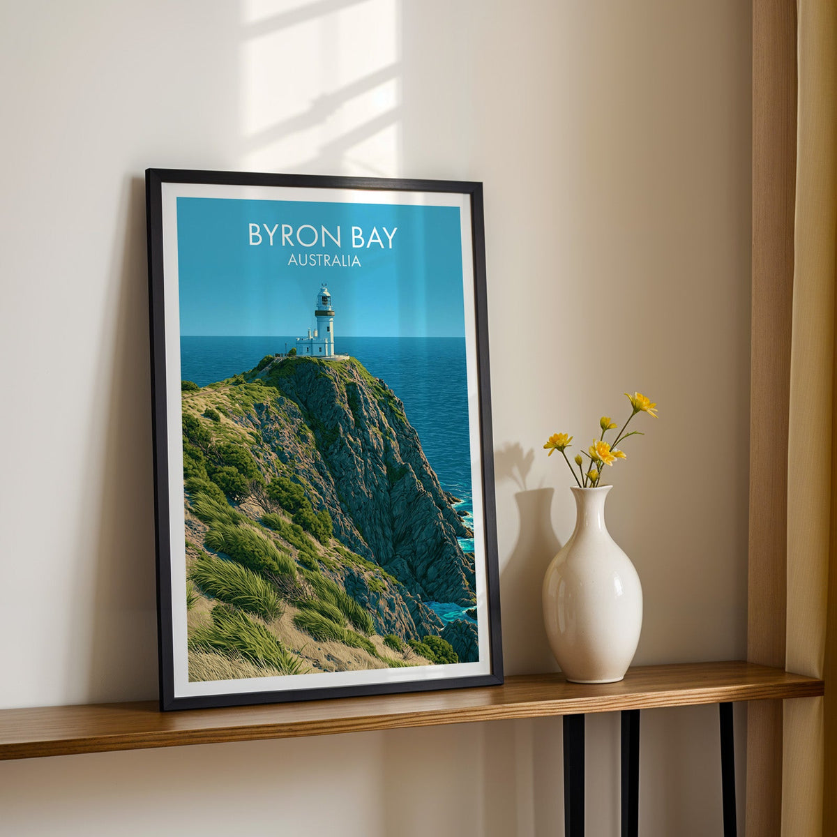 Cape Byron headland print in a black frame on a wood shelf with yellow daisies against a sunlit cream wall