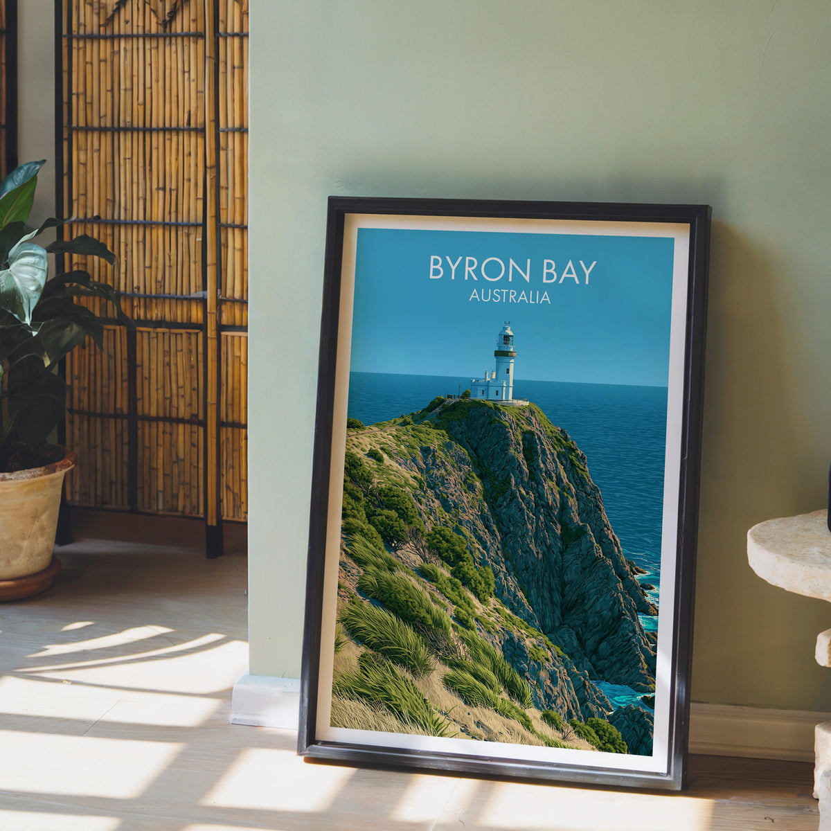 Byron Bay clifftop artwork in a black frame leaning on pale wood floor by a sage-green wall and bamboo screen