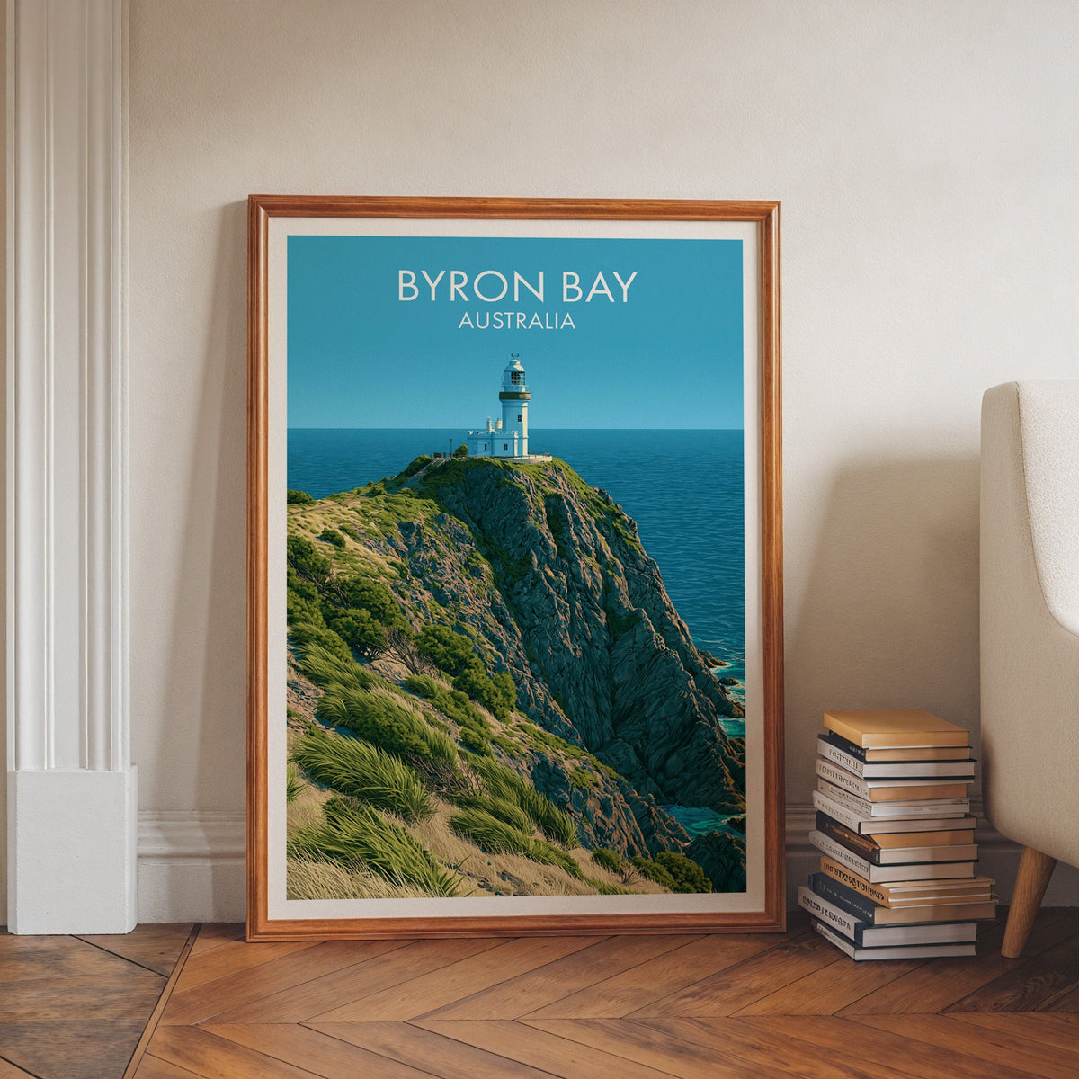 Lighthouse travel print in a walnut frame on herringbone floor beside stacked books and a pale armchair
