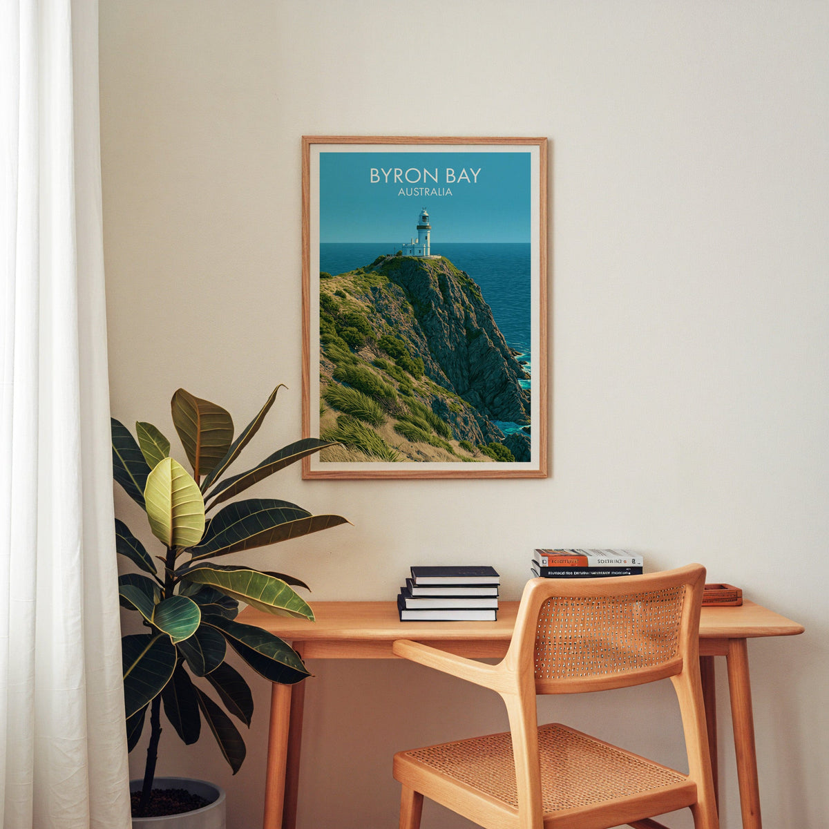 Cape Byron Lighthouse art in an oak frame above a wooden desk with a cane chair and rubber plant