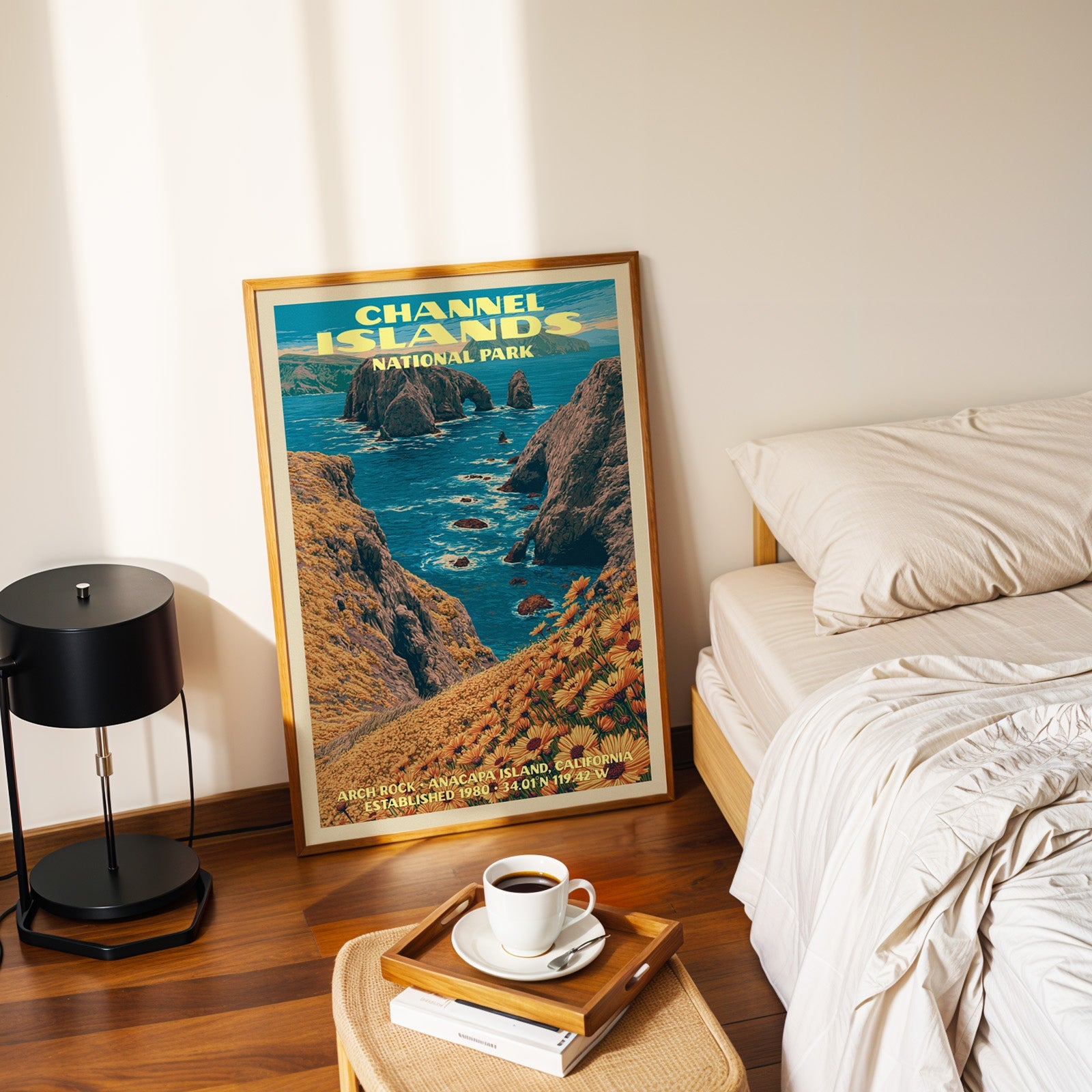 Arch Rock Channel Islands poster in a gold frame on the floor beside a made bed and a black table lamp