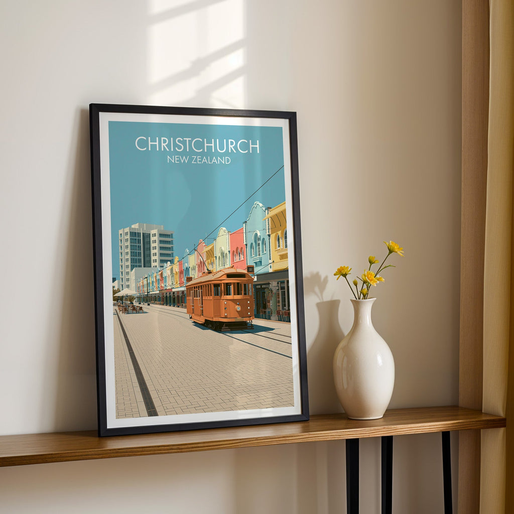 Orange tram and pastel shopfronts print, black frame on a wood shelf with daisies on a sunlit cream wall