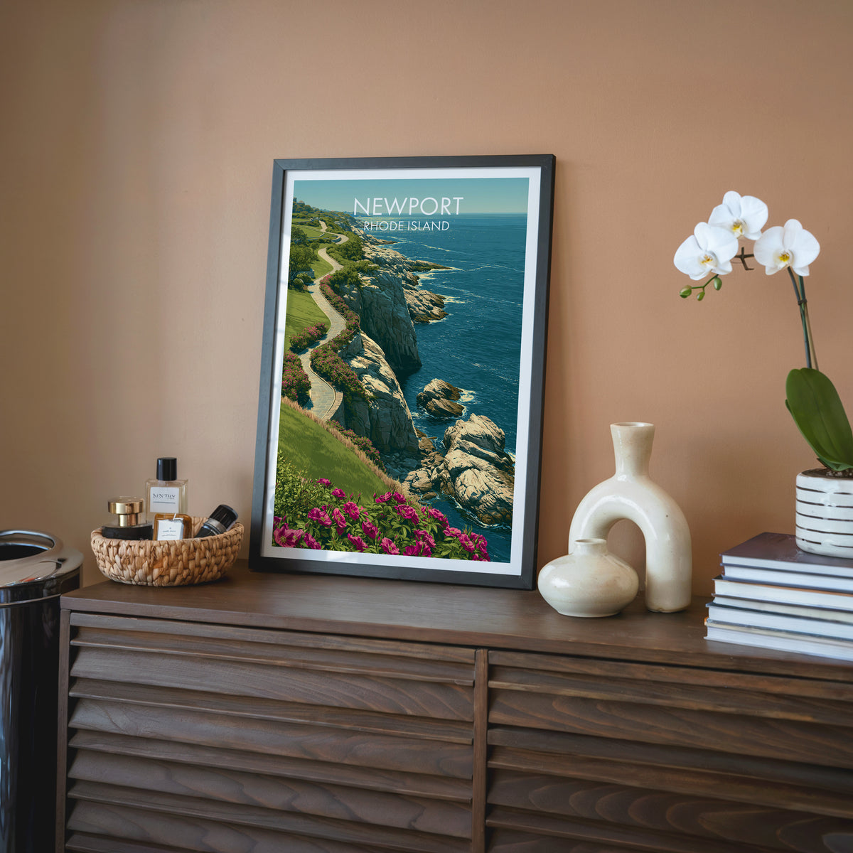 Coastal path wall art in a black frame on a walnut sideboard against a terracotta wall with an orchid