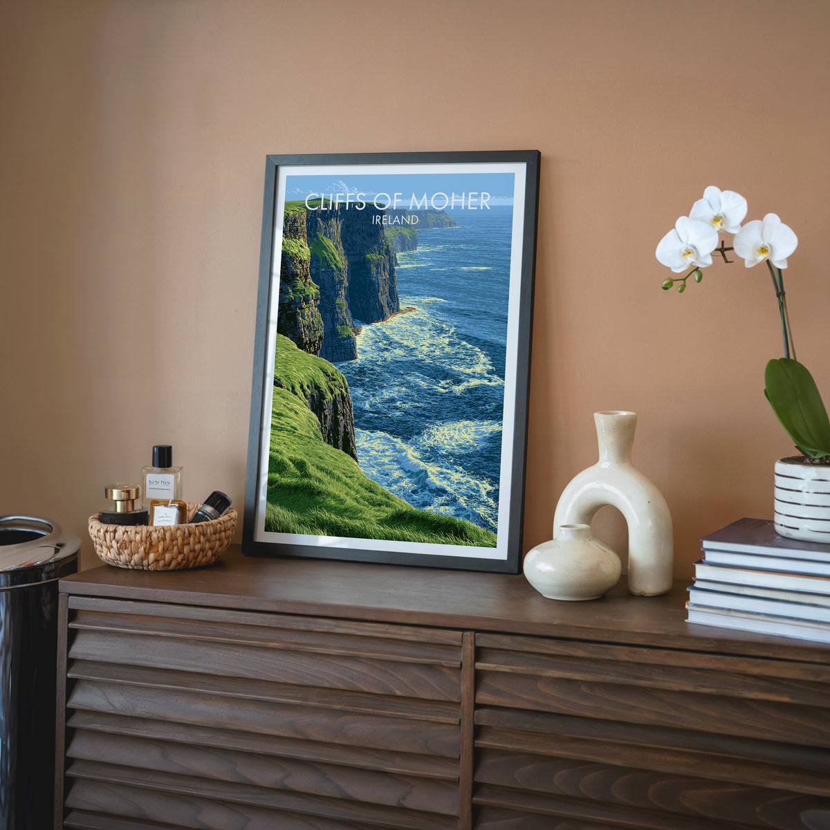 Ireland coastal wall art, black frame on a walnut sideboard against a terracotta wall with an orchid