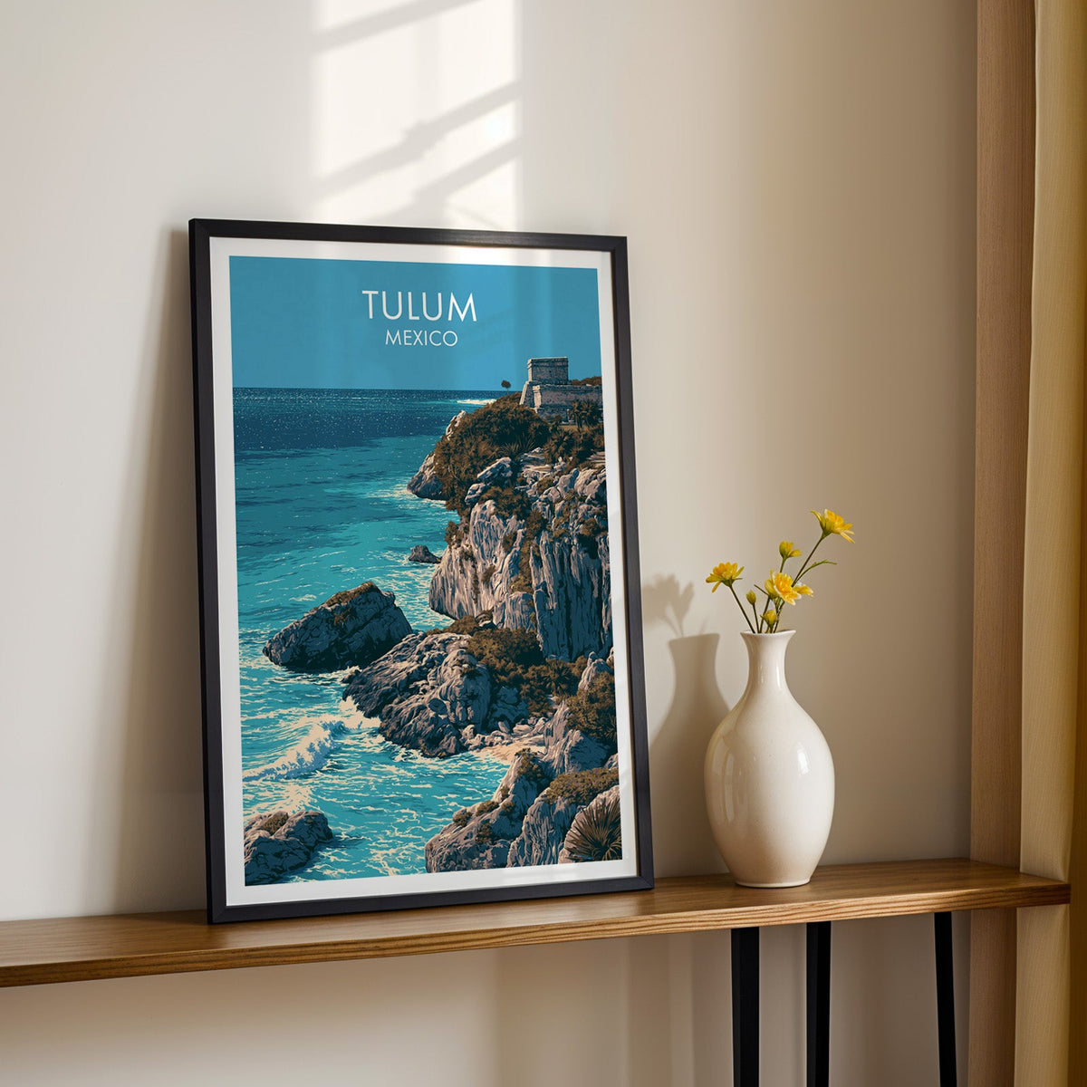 Maya ruin above the sea in a black frame on a wood shelf beside yellow daisies, sunlit cream wall.