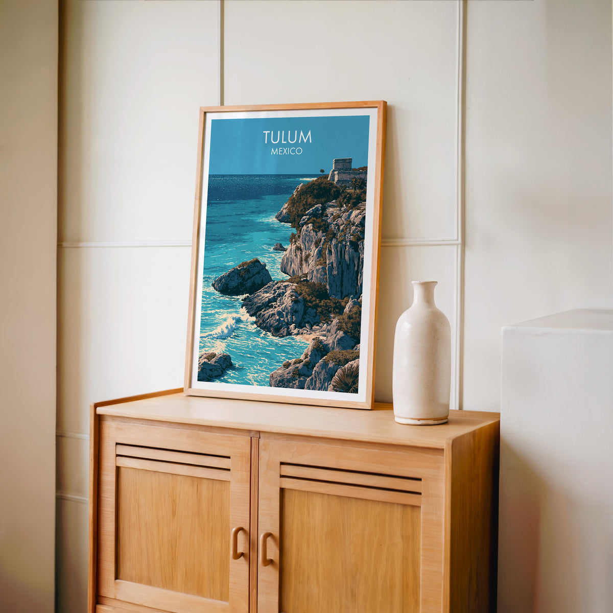 Navy and chalk grey coast art, oak frame on a light oak cabinet by a cream panelled wall and vase.