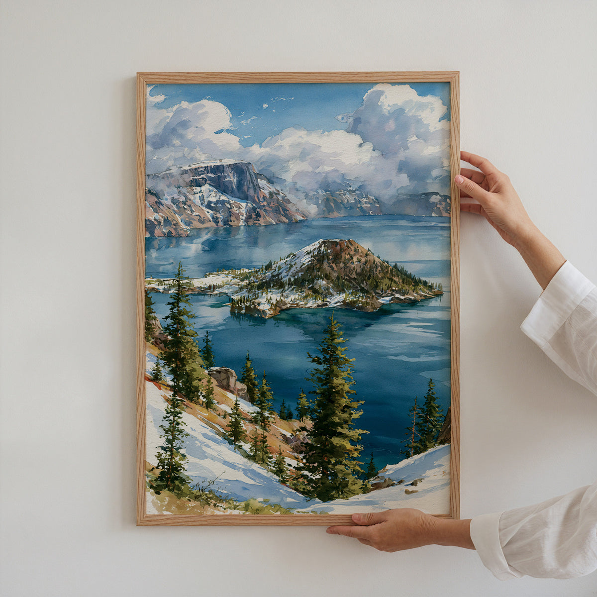 Hands holding an oak-framed Crater Lake National Park watercolor art print against a plain white wall