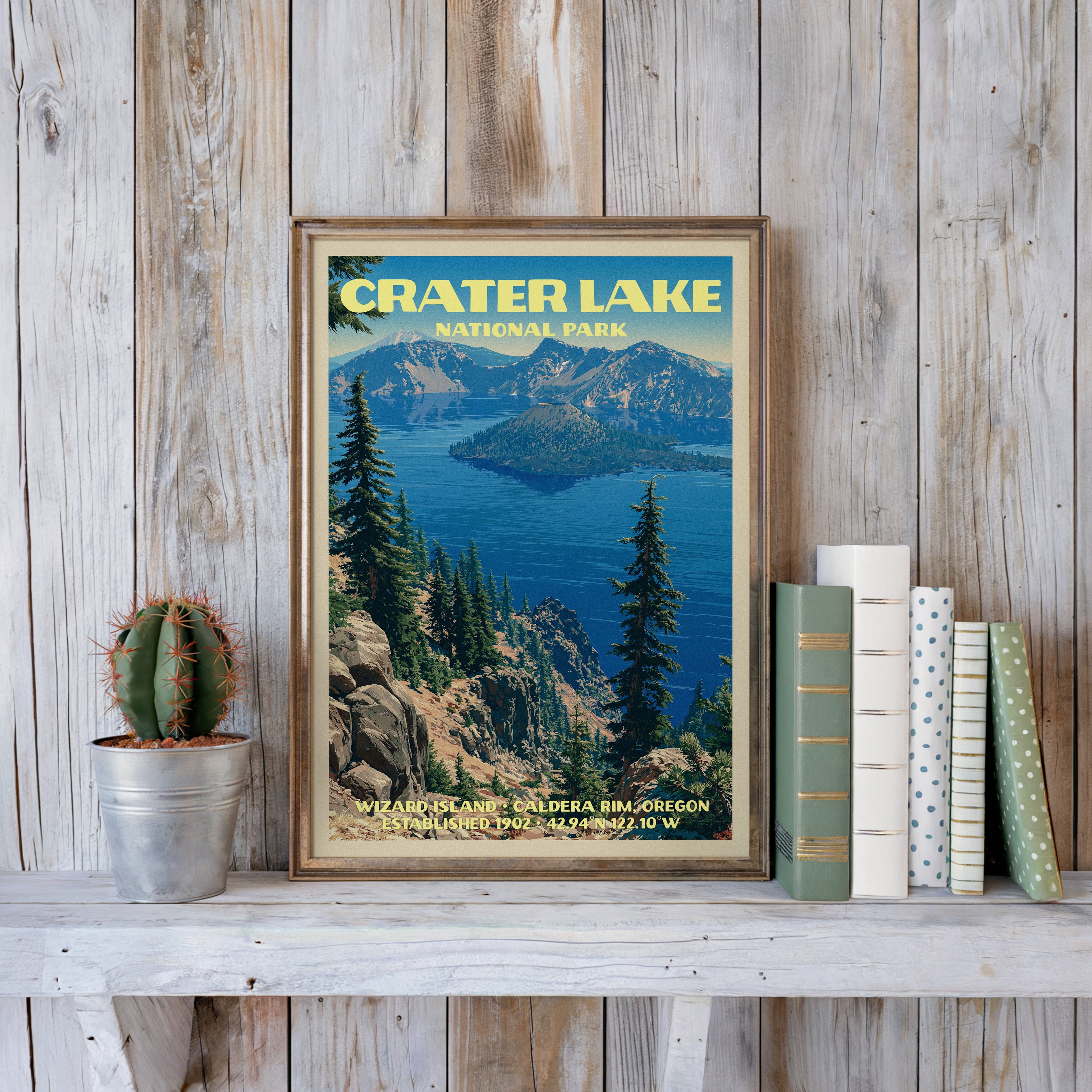 Crater Lake vintage poster in a slim gold frame on a whitewashed plank wall beside a cactus and pastel books