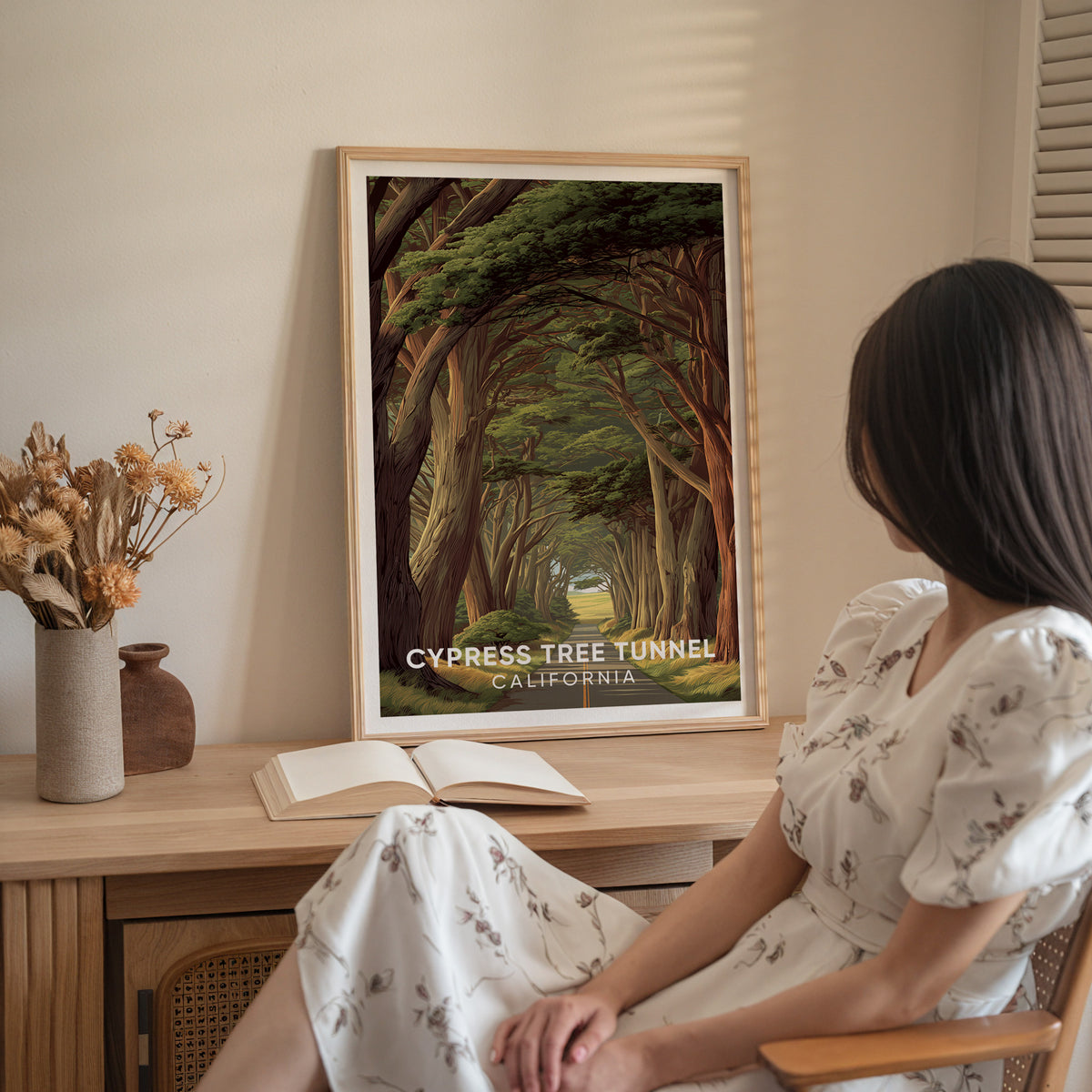 Woman in a white dress seated beside an oak-framed cypress tunnel print on a wooden sideboard.