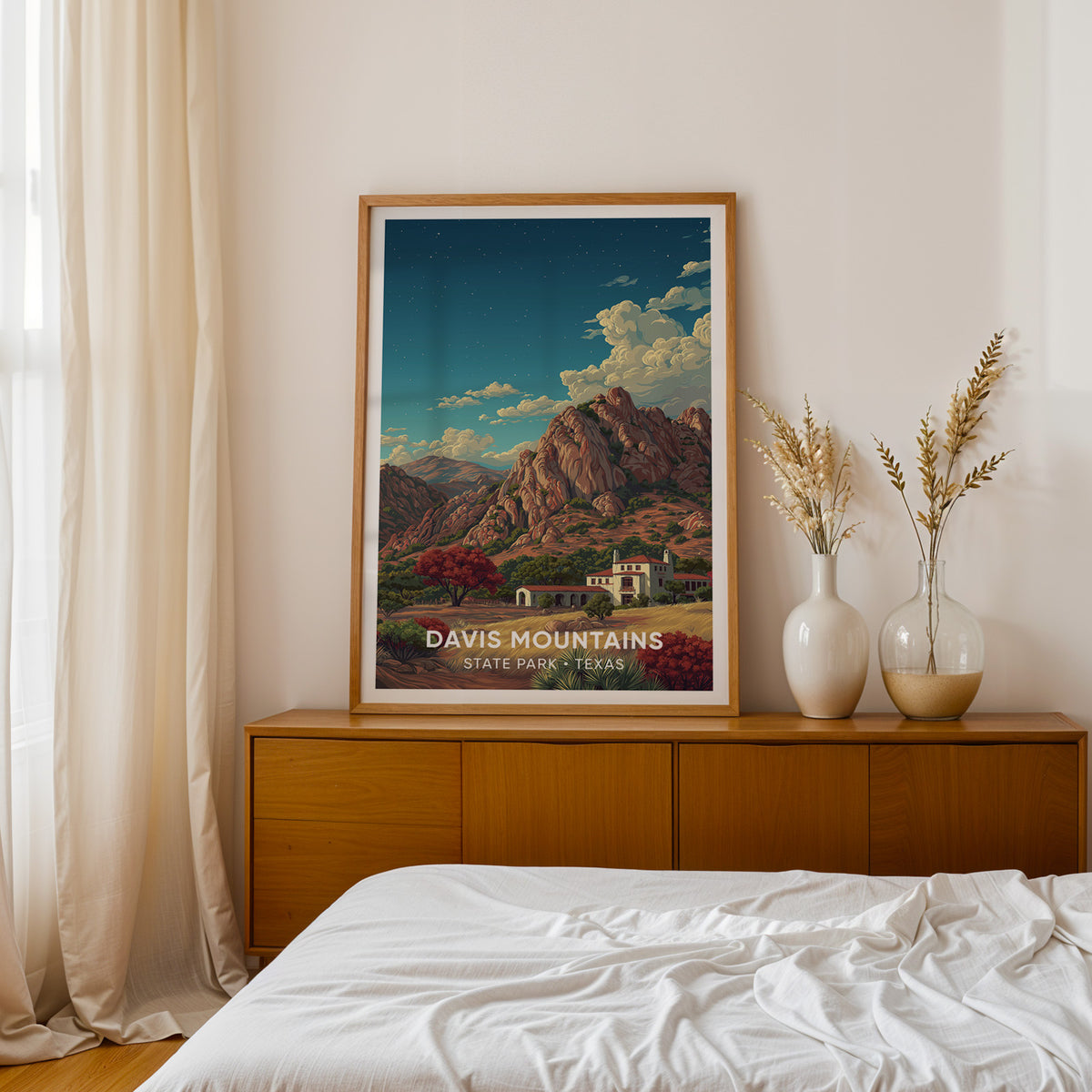 Davis Mountains print in an oak frame on a teak sideboard beside a bed, dried pampas grass in two white vases.