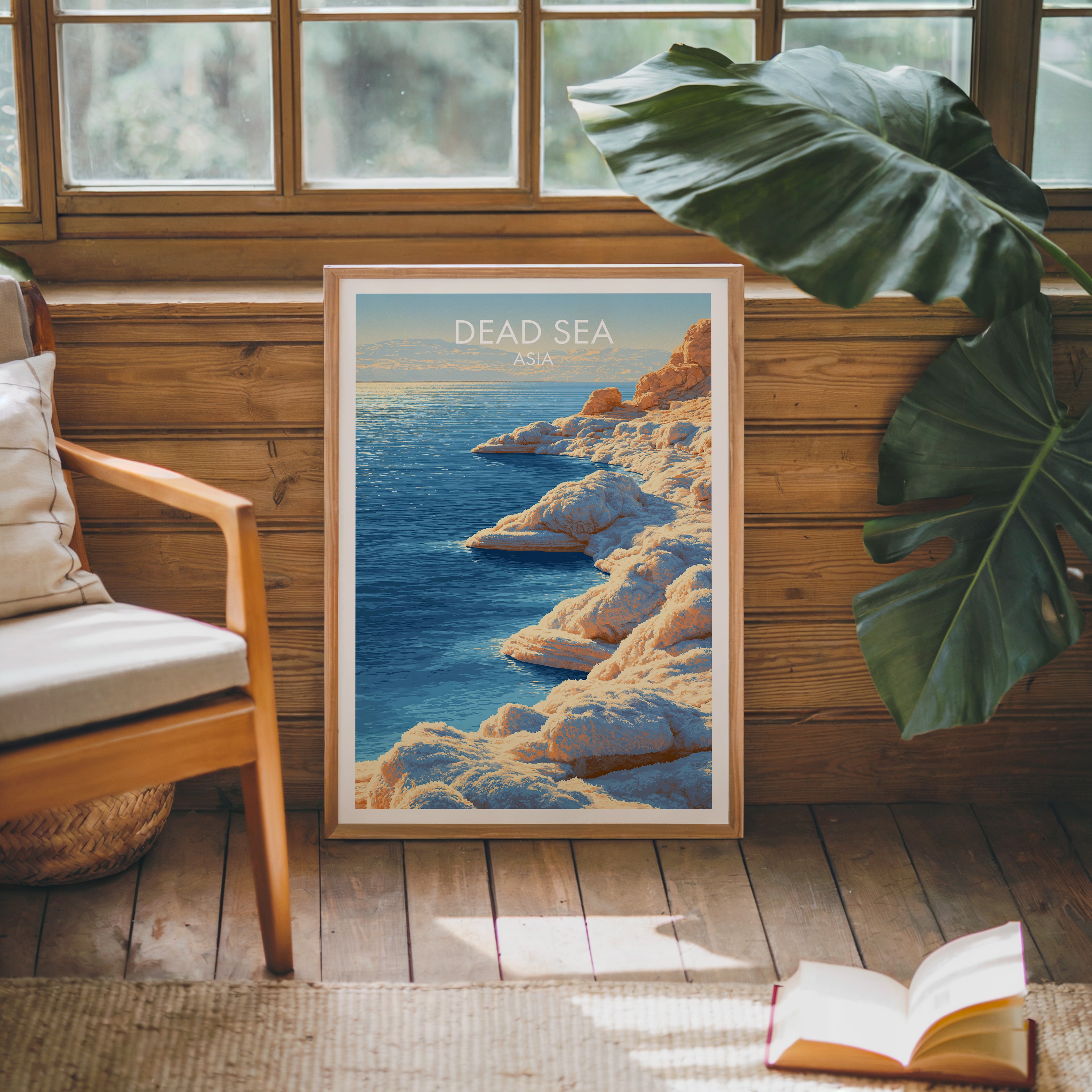 Framed Dead Sea Middle East poster leaning against a timber cabin wall with monstera leaves and an open book