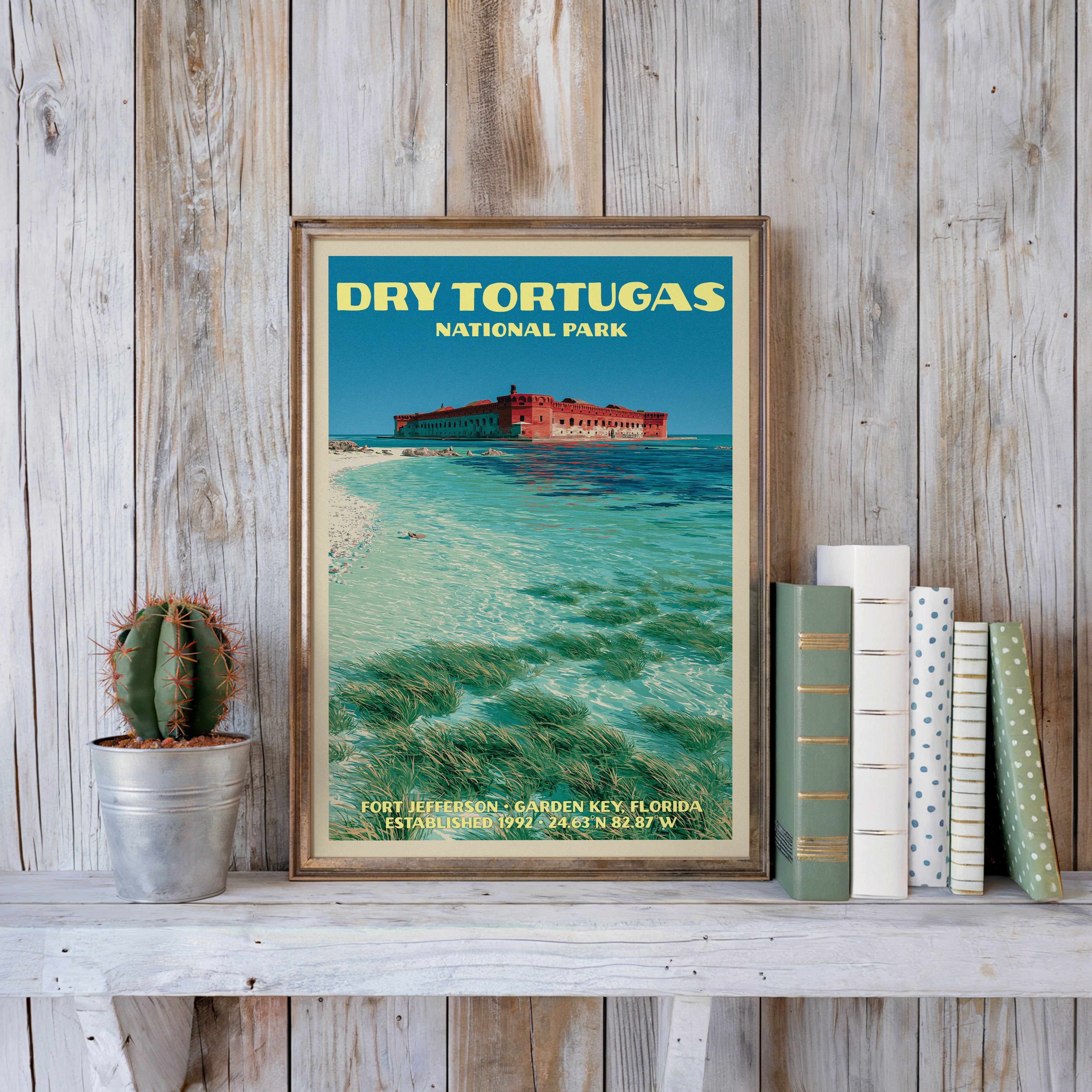 Dry Tortugas vintage poster in a slim gold frame on a whitewashed plank wall beside a cactus and pastel books