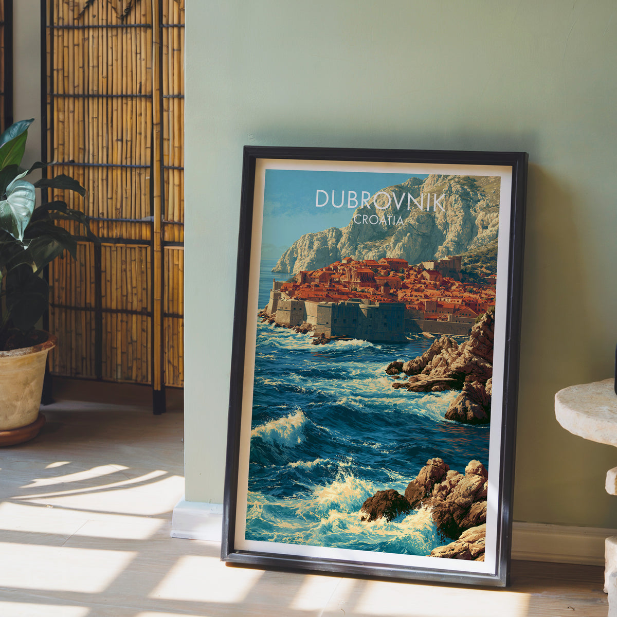 Dubrovnik walls poster, dark frame leaning on a sage-green wall beside a bamboo screen and a fern