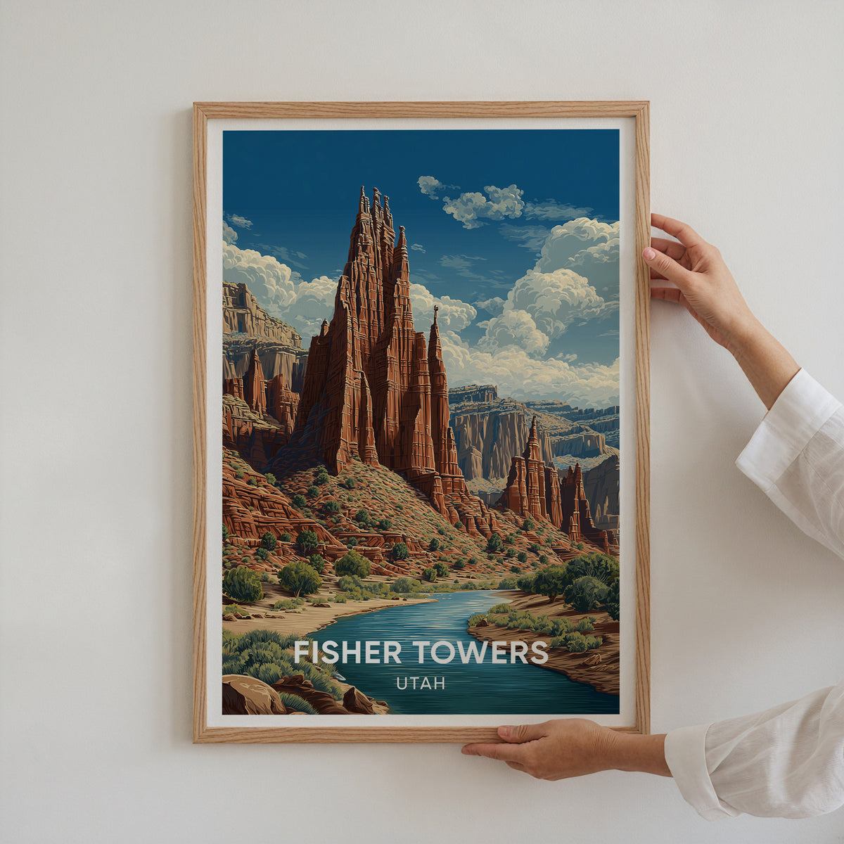 Two hands holding the Fisher Towers poster in an oak frame up against a plain white wall.