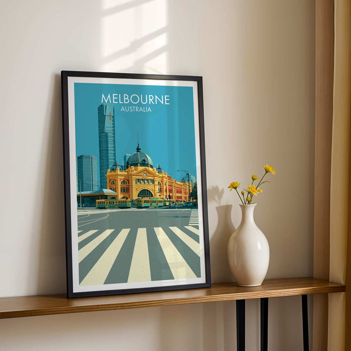 Flinders Street artwork in a black frame on a wood shelf beside a vase of yellow daisies, sunlit cream wall