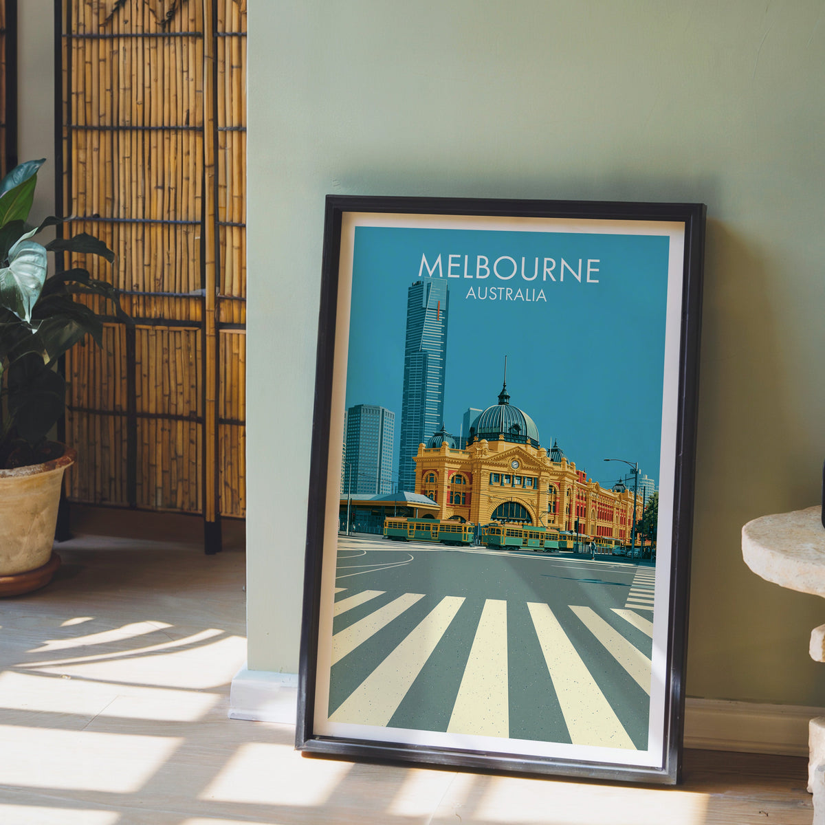Melbourne tram scene print in a black frame leaning on pale wood floor by a sage green wall and bamboo screen