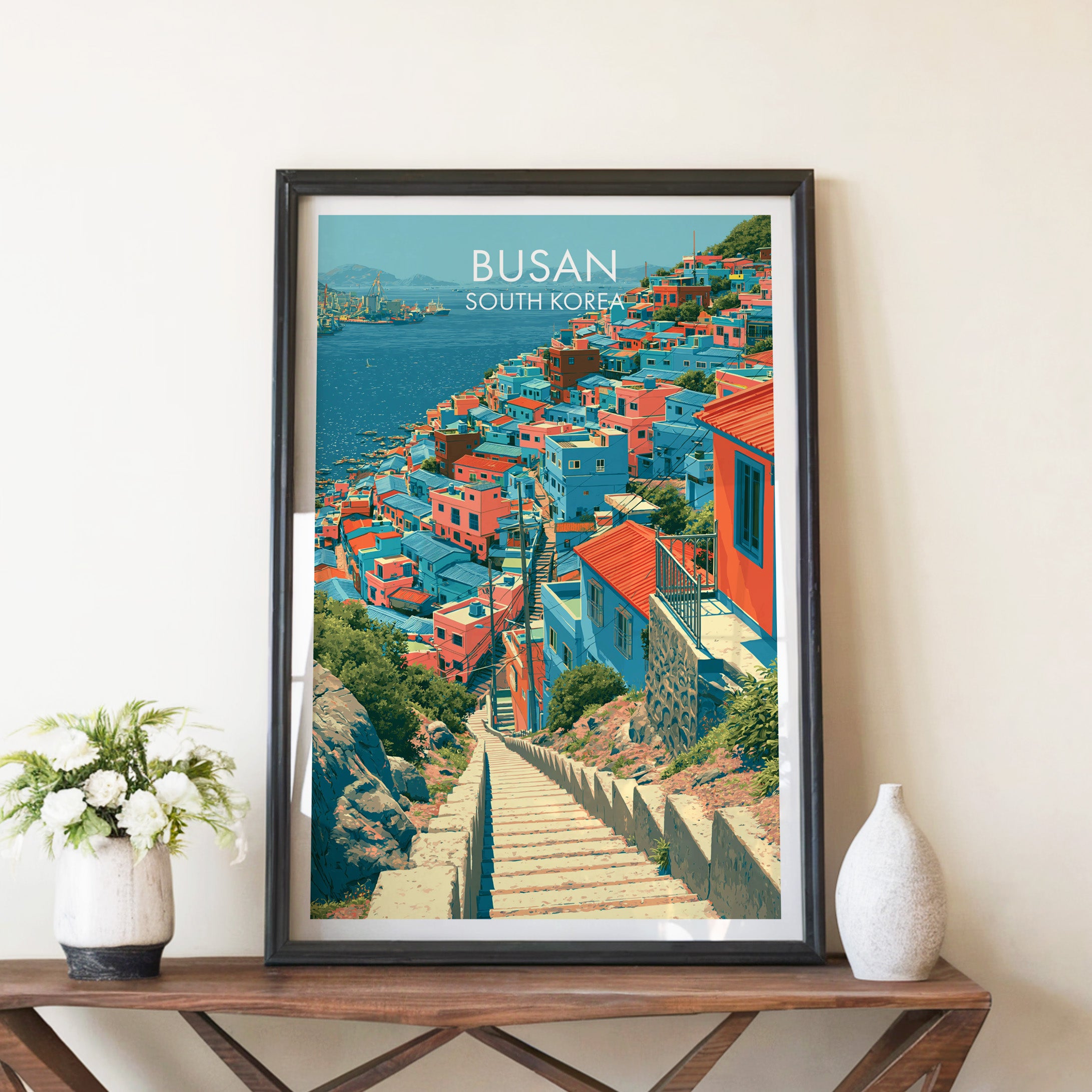 Gamcheon Culture Village Busan travel poster in a black frame on a wooden console table with white flowers