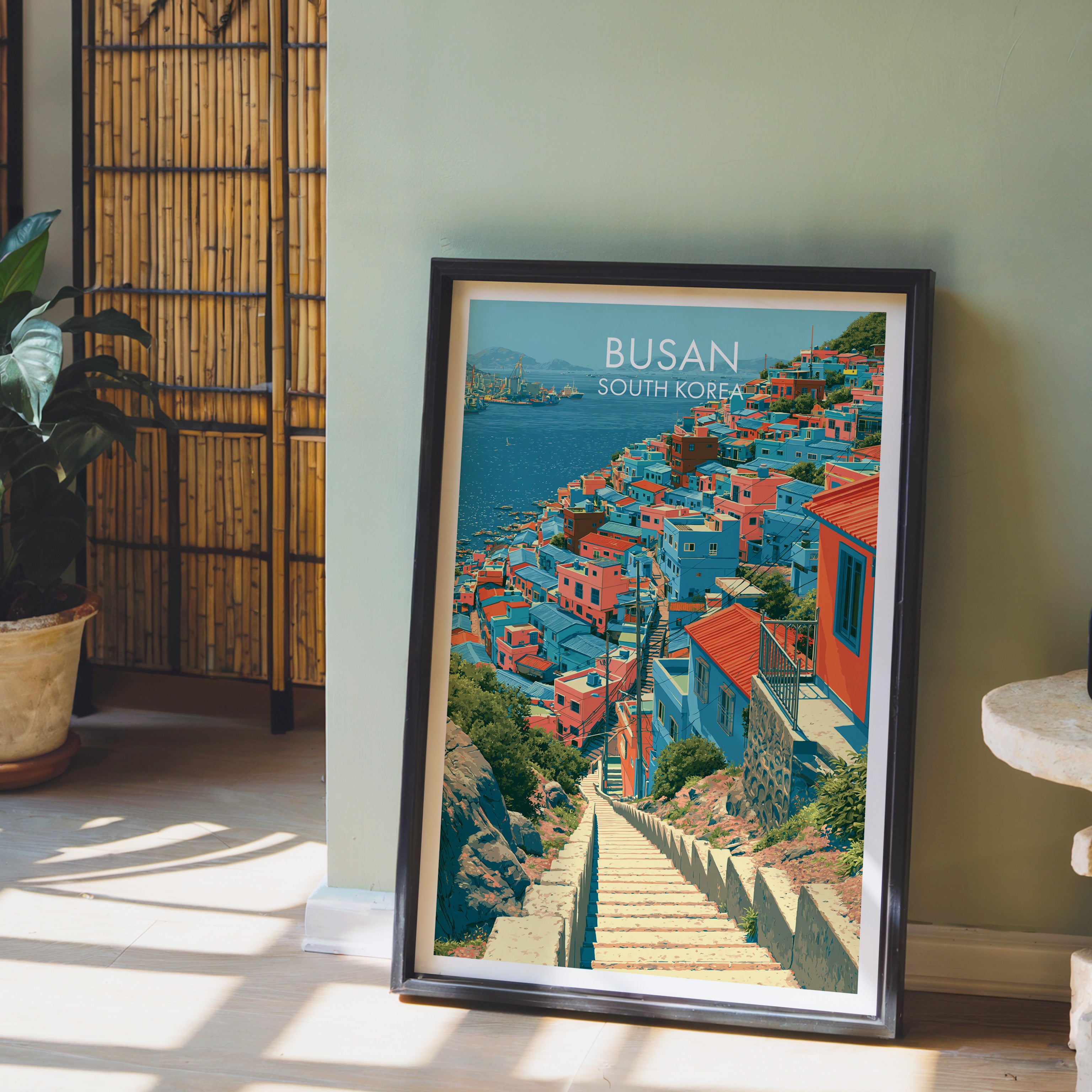 Gamcheon Culture Village Busan wall art in a black frame leaning against a sage green wall beside a bamboo screen
