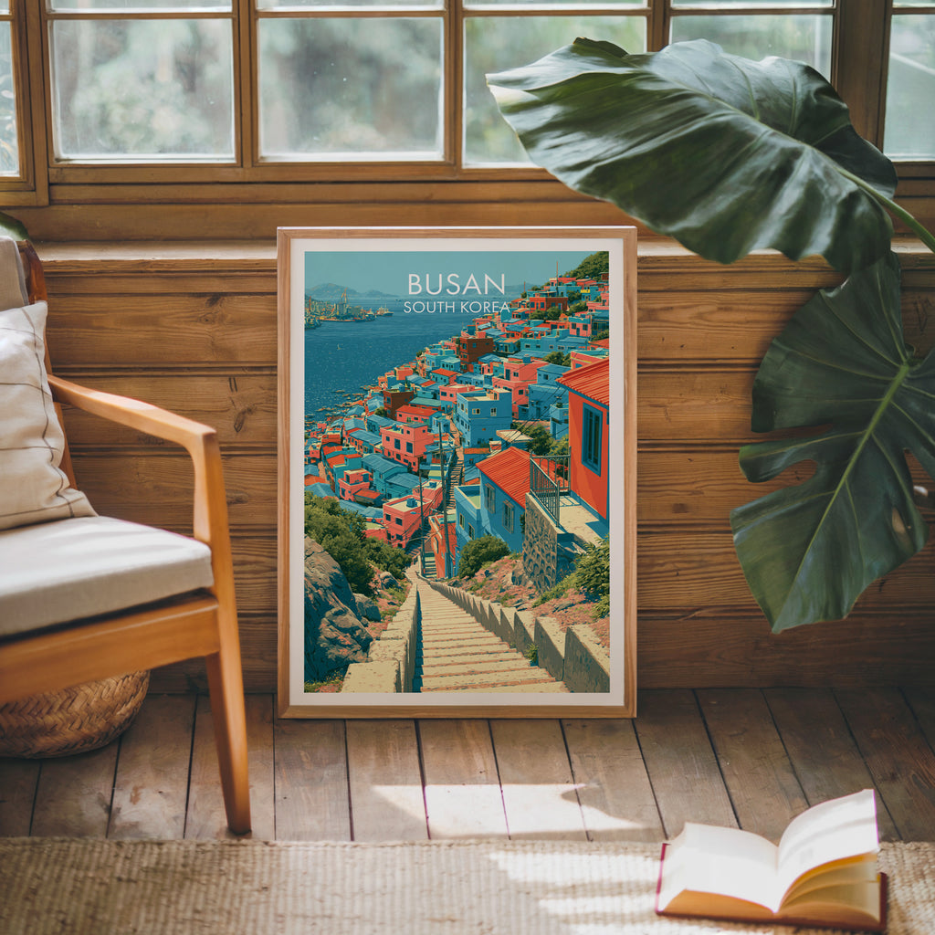 Framed Gamcheon Culture Village Busan poster leaning against a timber cabin wall with monstera leaves and an open book