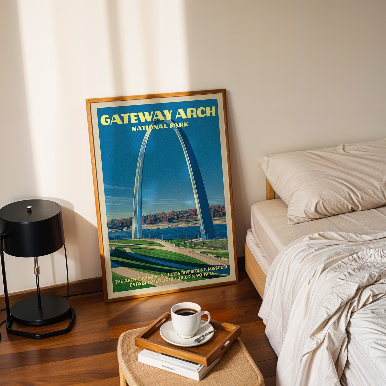 St. Louis riverfront Gateway Arch poster in a gold frame on the floor beside a made bed and a table lamp