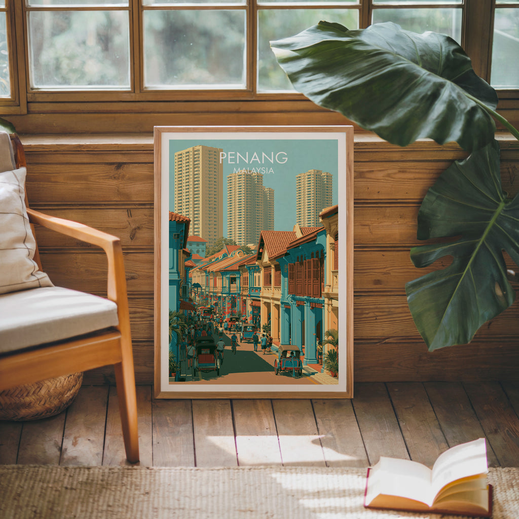 Framed Georgetown Penang Malaysia poster leaning against a timber cabin wall with monstera leaves and an open book