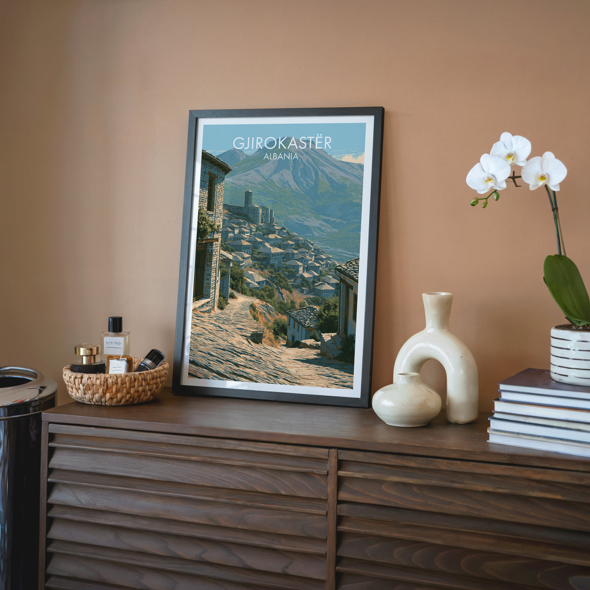 Gjirokaster Albania wall art, black frame on a walnut sideboard against a terracotta wall with a white orchid