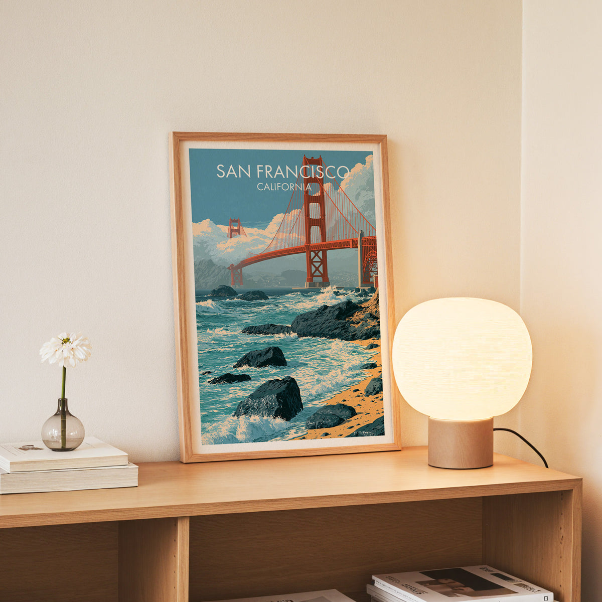 Golden Gate artwork in a slim oak frame standing on a console with daisies in a glass jar and books.