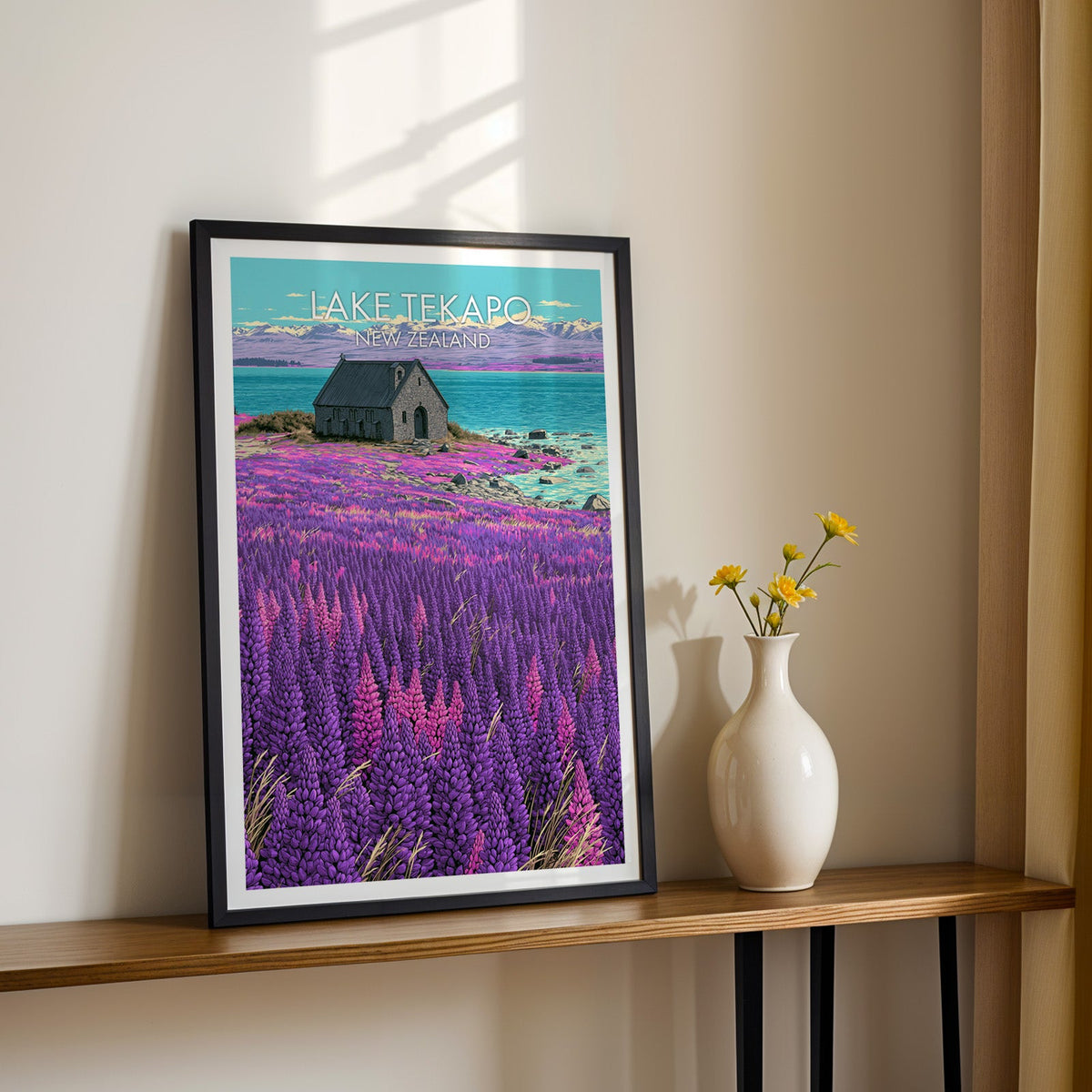 Stone chapel artwork in a black frame on a wood shelf with yellow daisies on a sunlit cream wall