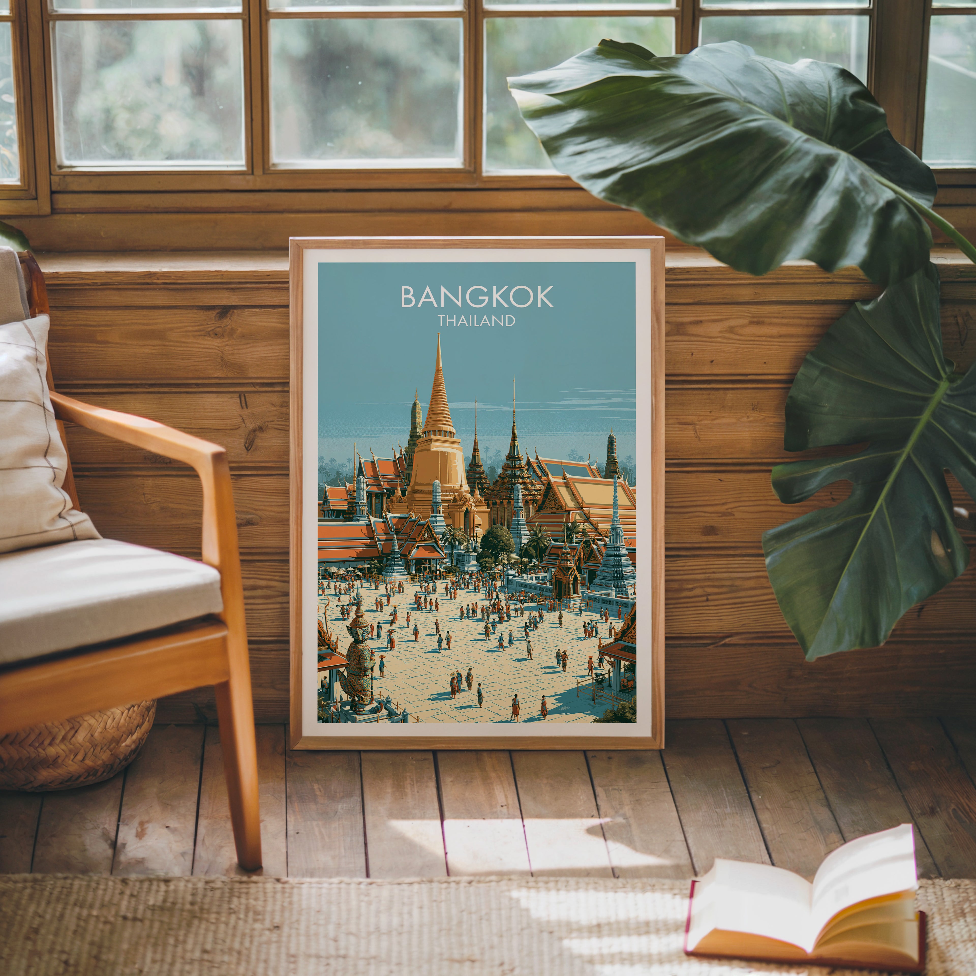 Framed Grand Palace Bangkok Thailand poster leaning against a timber cabin wall with monstera leaves and an open book