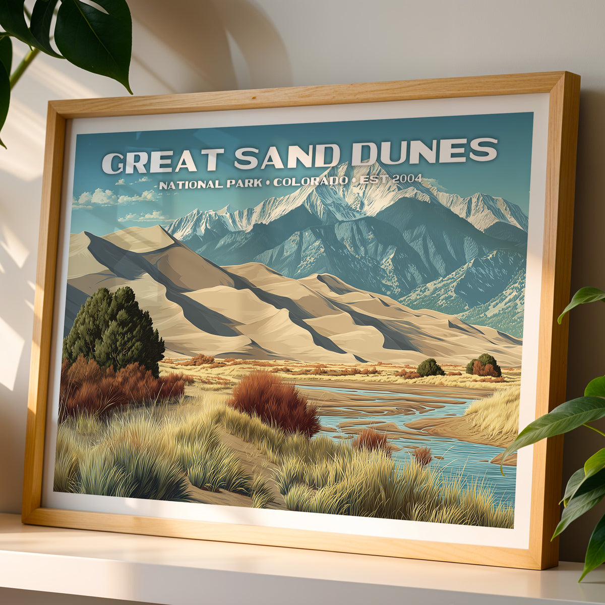 Great Sand Dunes National Park wall art print in a light oak frame, leaning on a white shelf in warm sunlight beside a monstera plant