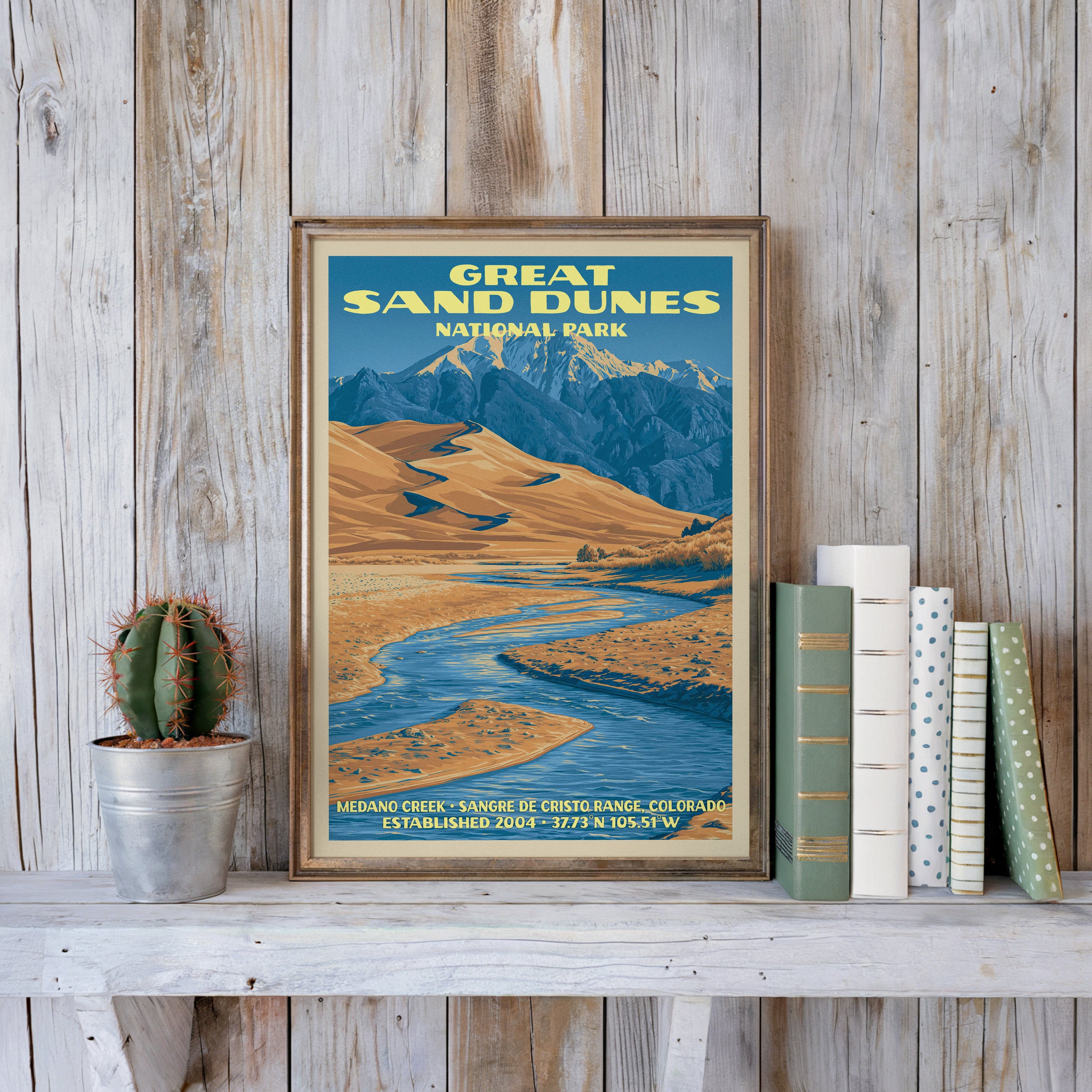 Medano Creek poster in a slim gold frame on a whitewashed plank wall beside a tin-potted cactus and pastel books