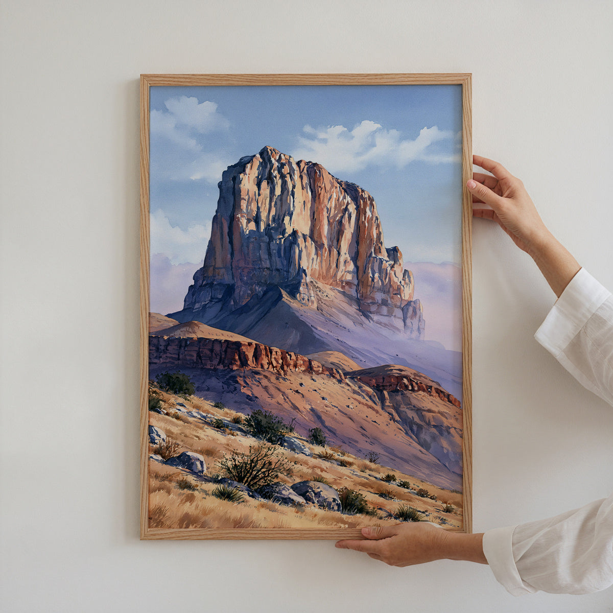 Hands holding an oak-framed Guadalupe Mountains watercolor art print against a plain white wall