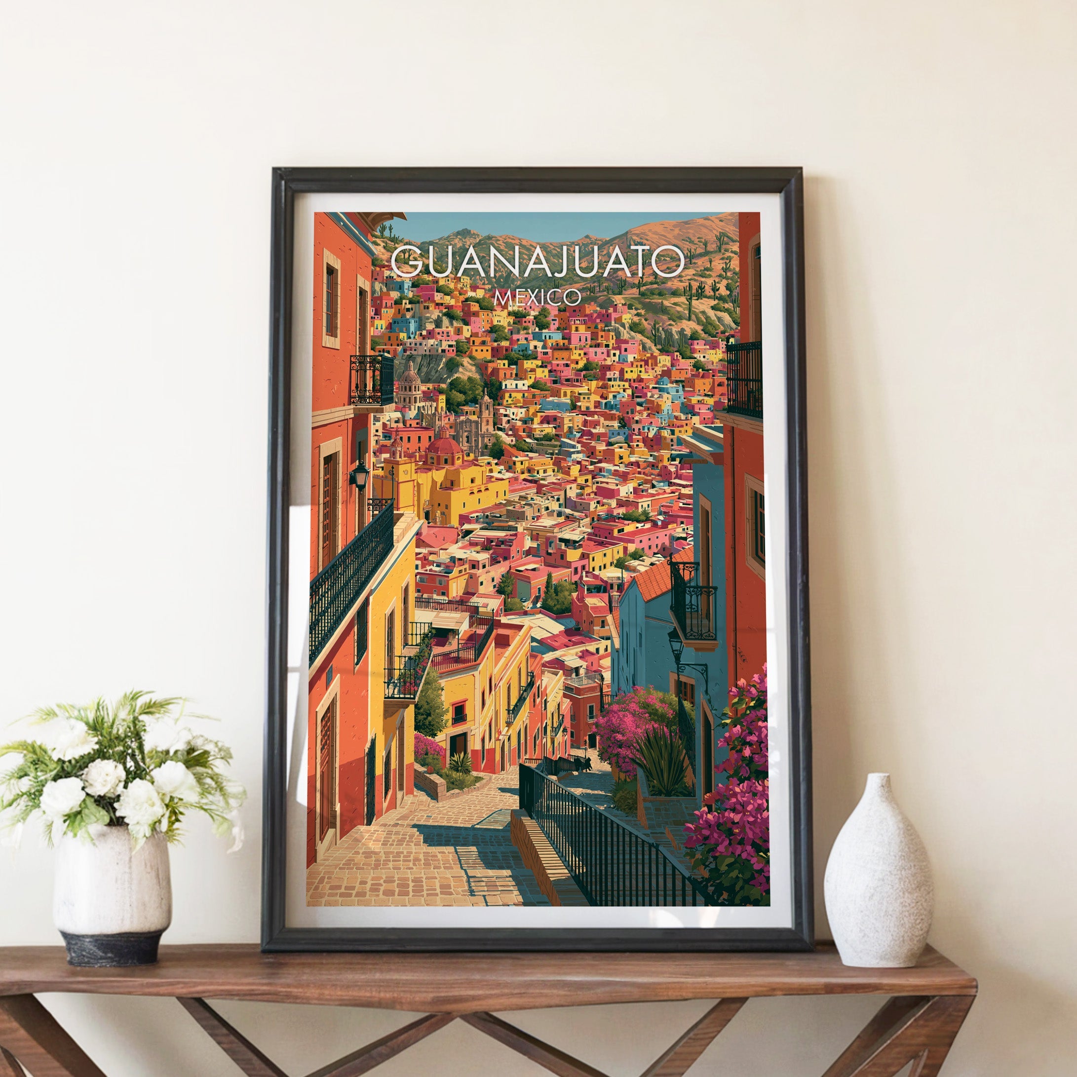 Colourful Mexican hill town poster in a black frame on a walnut console with white peonies and a ceramic vase