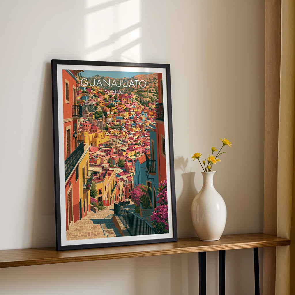 Cobbled alley view of Guanajuato in a black frame on a wood shelf with yellow daisies, sunlit cream wall