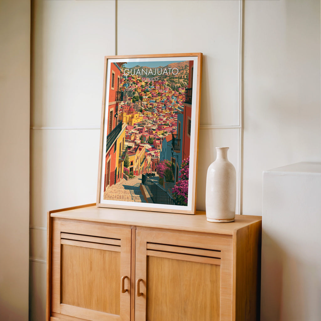 Hill town artwork in an oak frame on a light oak cabinet against a cream panelled wall with a tall vase